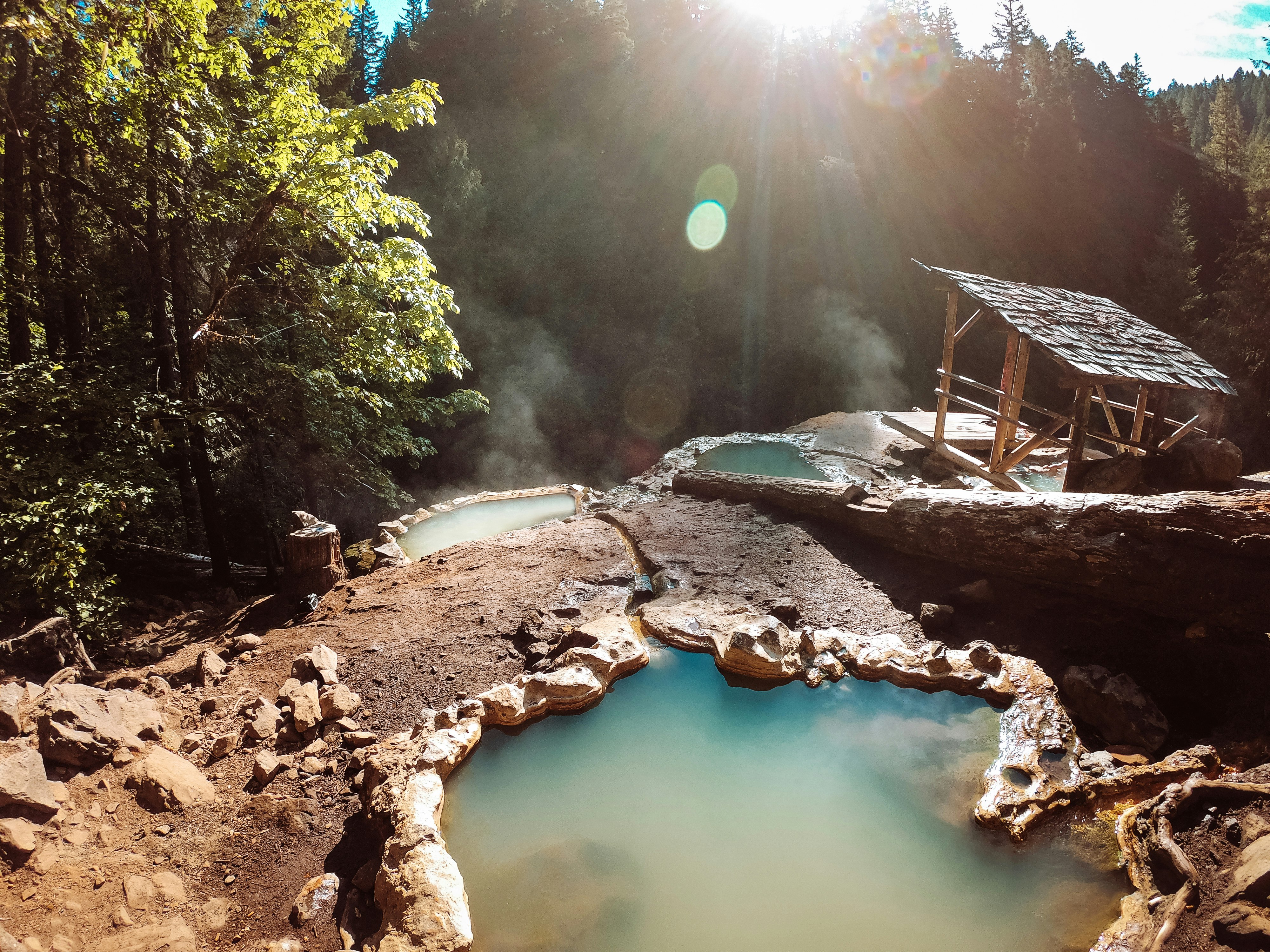 Three deep irregularly shaped teal pools surrounded by light brown earth and rocks overlook a stand of spruce and fir trees. A lens flare cuts through on the right side of the frame, highlighting a small wooden shelter with a wooden shingle roof covering one of the pools.