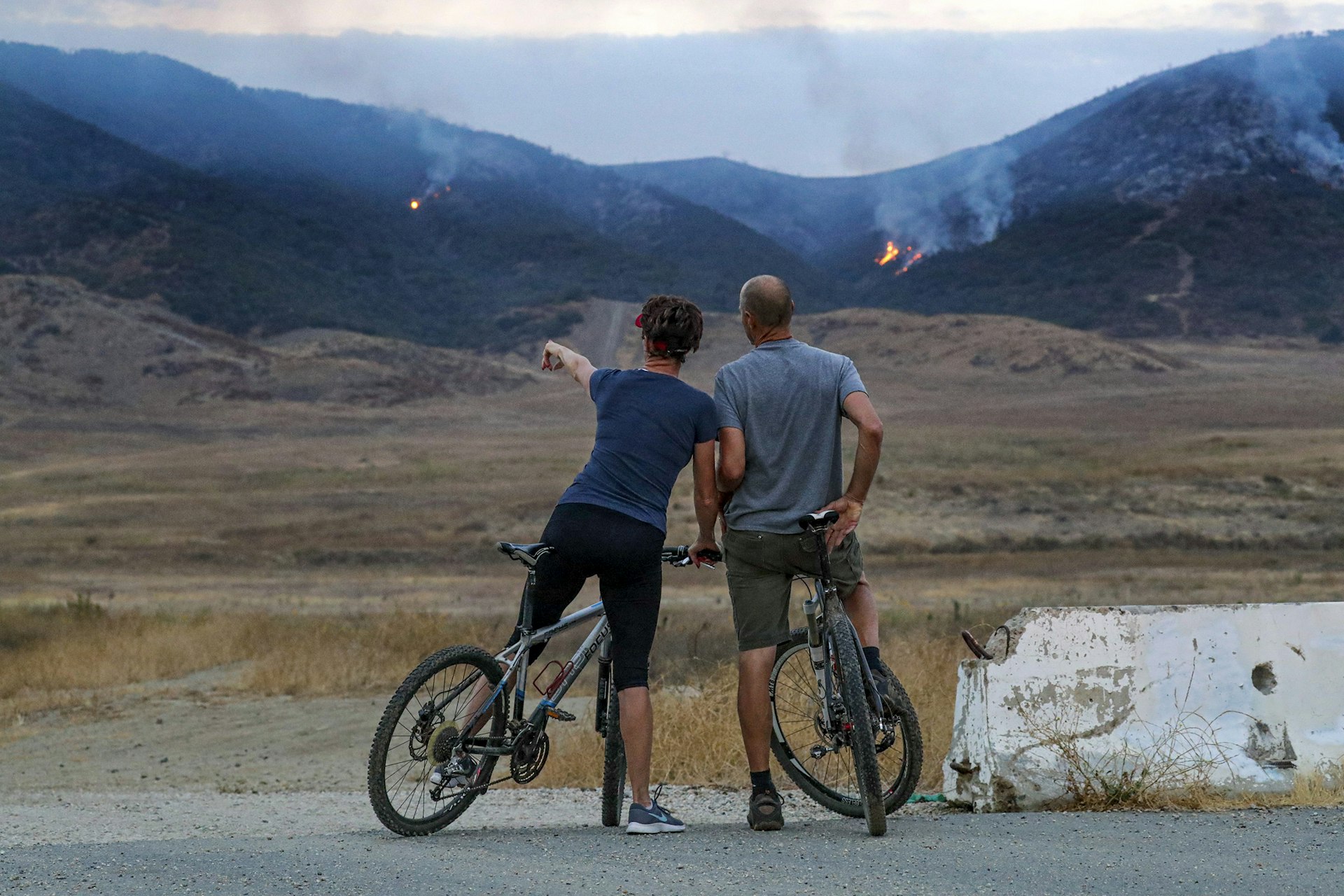 a man and a woman stand with their bikes pointing to small fires burning in the mountains in the distance
