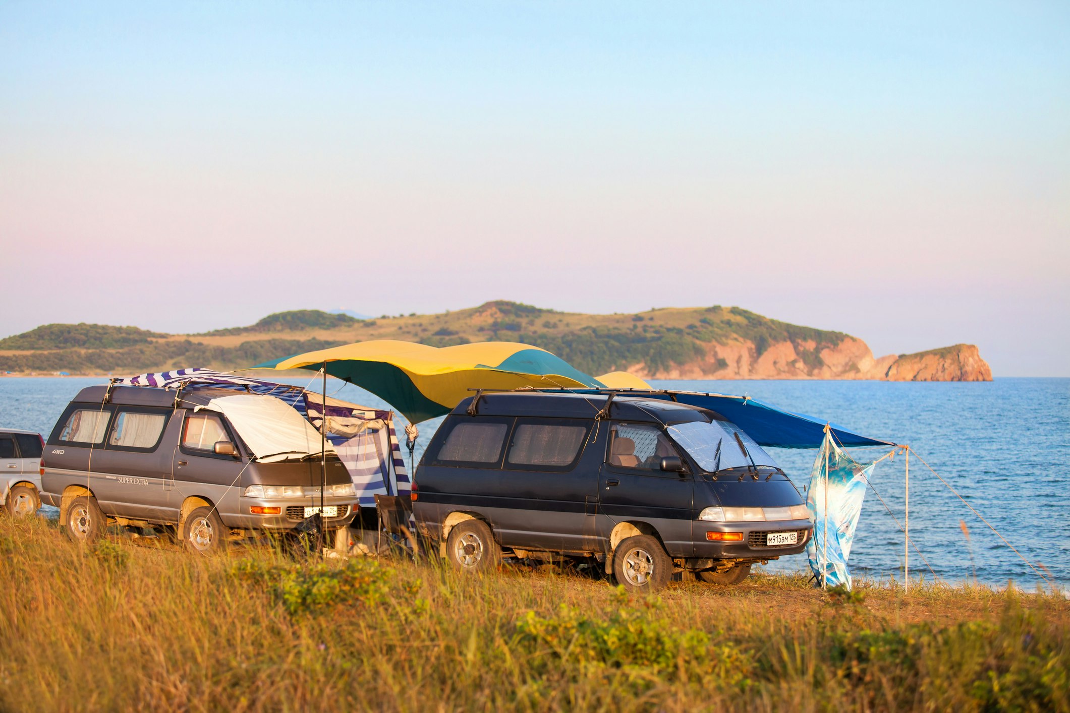 Two Toyota vans are parked by the seashore, with yellow and blue tarps and pop-up canopies to make the impromptu campsite more comfortable.