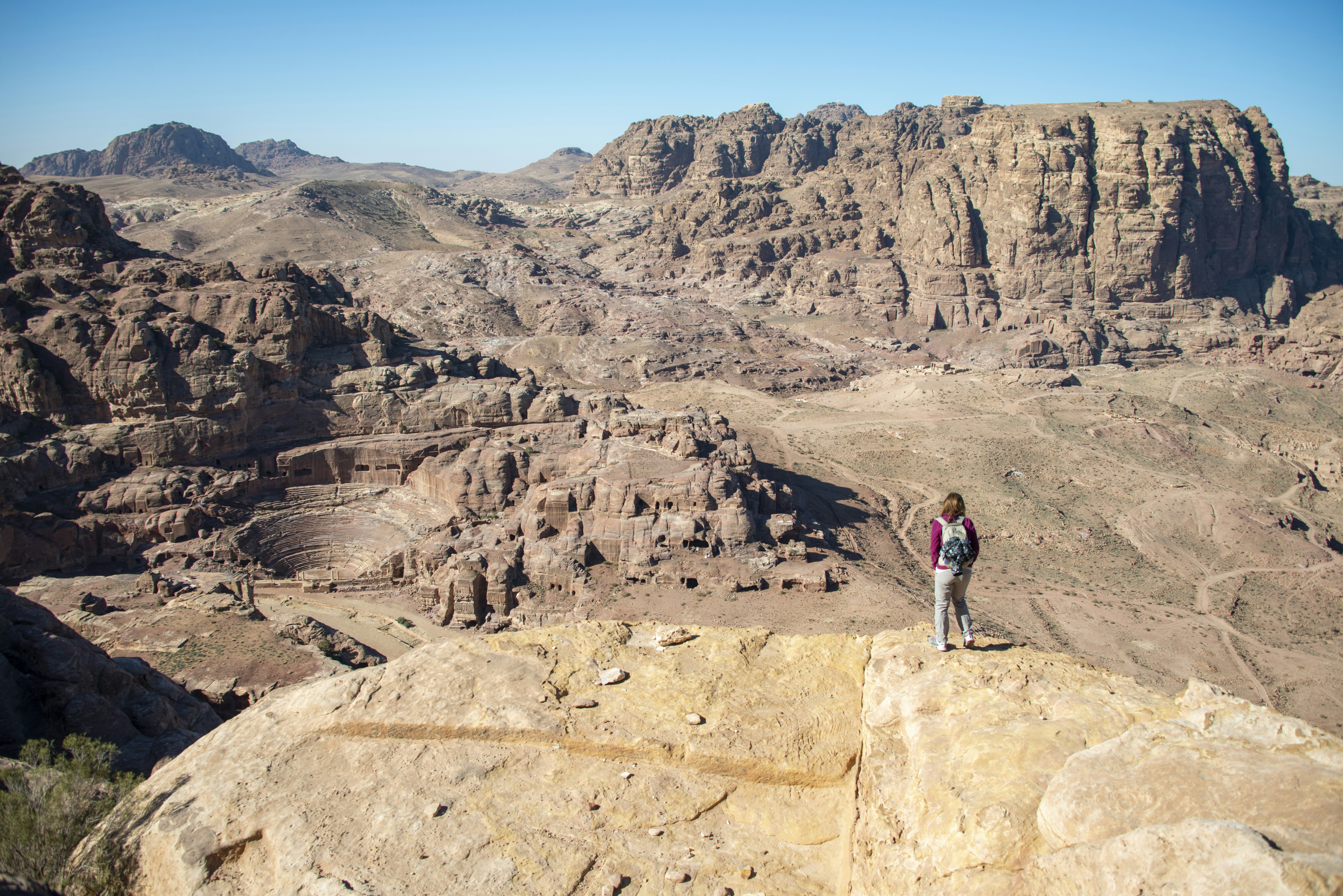 A woman in grey pants and a pink shirt with a grey backpack stands on a rocky overlook with an expansive view of the Nabatean theater in the Petra valley along the Jordan Trail