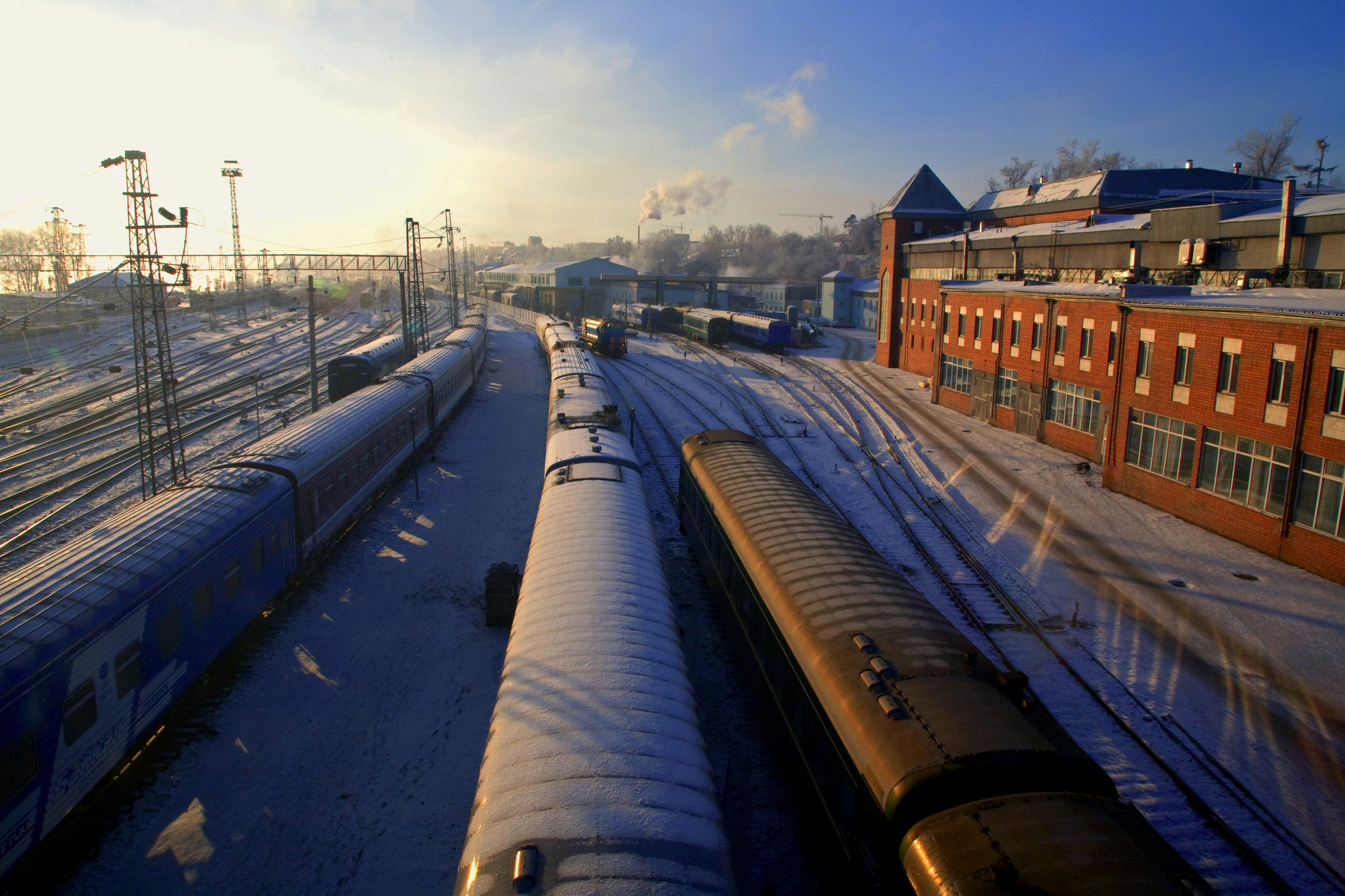 Three trains lined up on the tracks next to a red-brick train station. Snow lies on the grounds and between the tracks, and the sun is low in the sky.