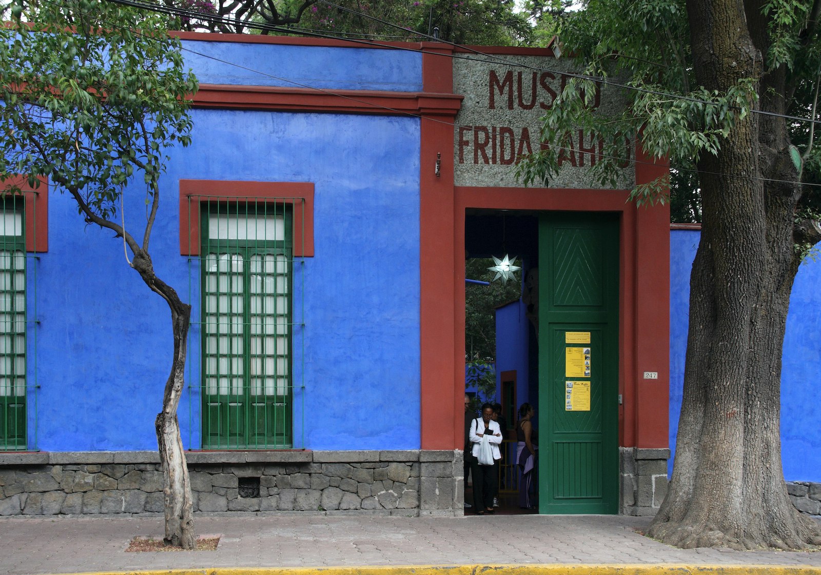 The Frida Kahlo museum- a blue concrete building with red trip, green windows, a green door, and a tree in guarding the front door