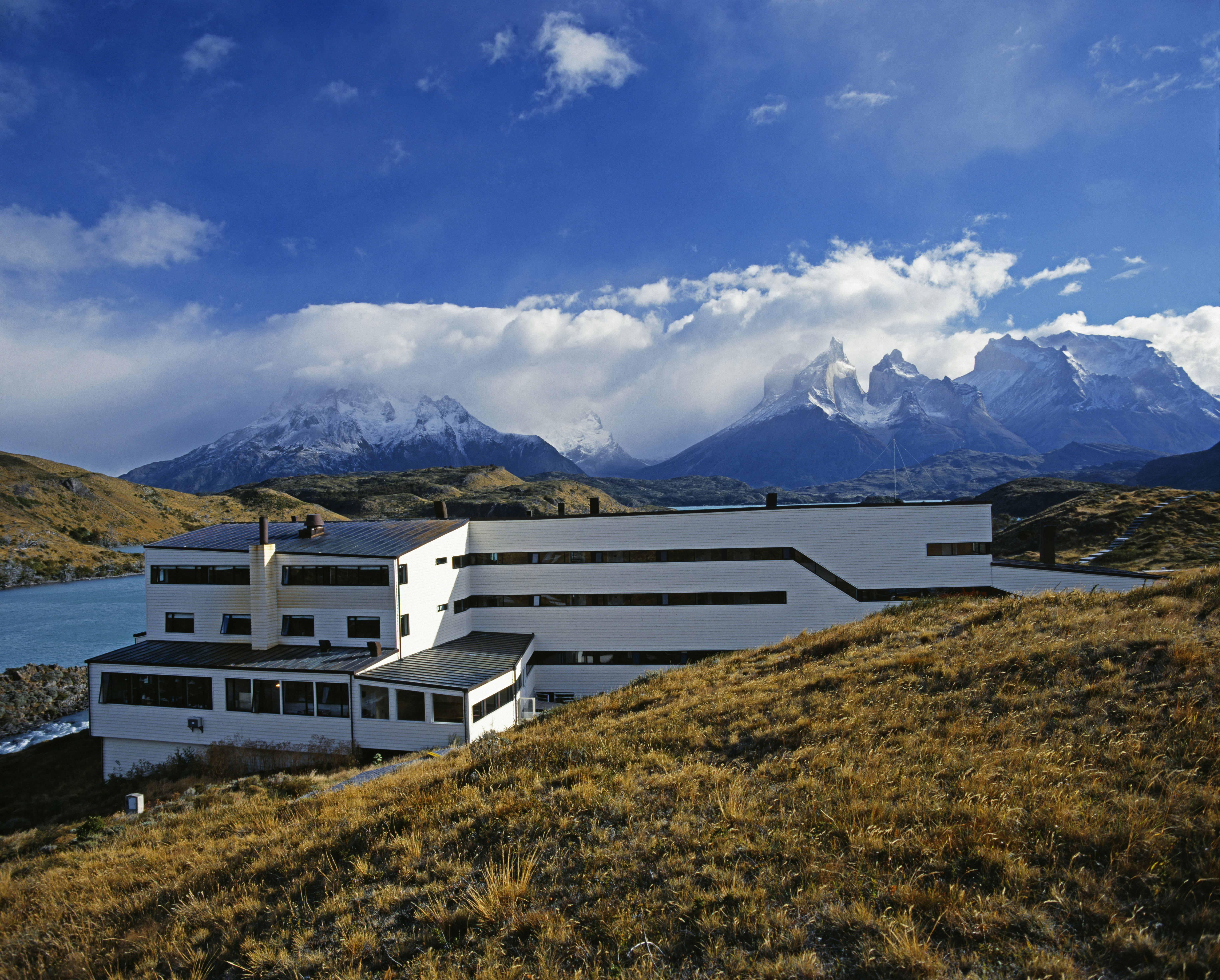 The white, modernist form of the Explora Hotel in Salto Chico Patagonia is set into a hillside covered in greenish yellow scrub. In the distance, you see the Paine Massif and a glacial lake which the hotel overlooks, under a deep blue sky and white lines of clouds.