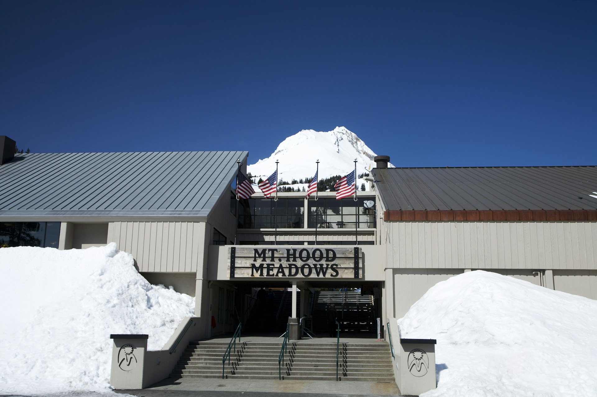 The snowy peak of Mt. Hood is framed by the entrance to Mt. Hood Meadows ski resort, which is made up of a large serif-font sign with the name of the lodge balanced over the front doors between two plain A-Frame wings of the building. Three American flags flutter over the signage.