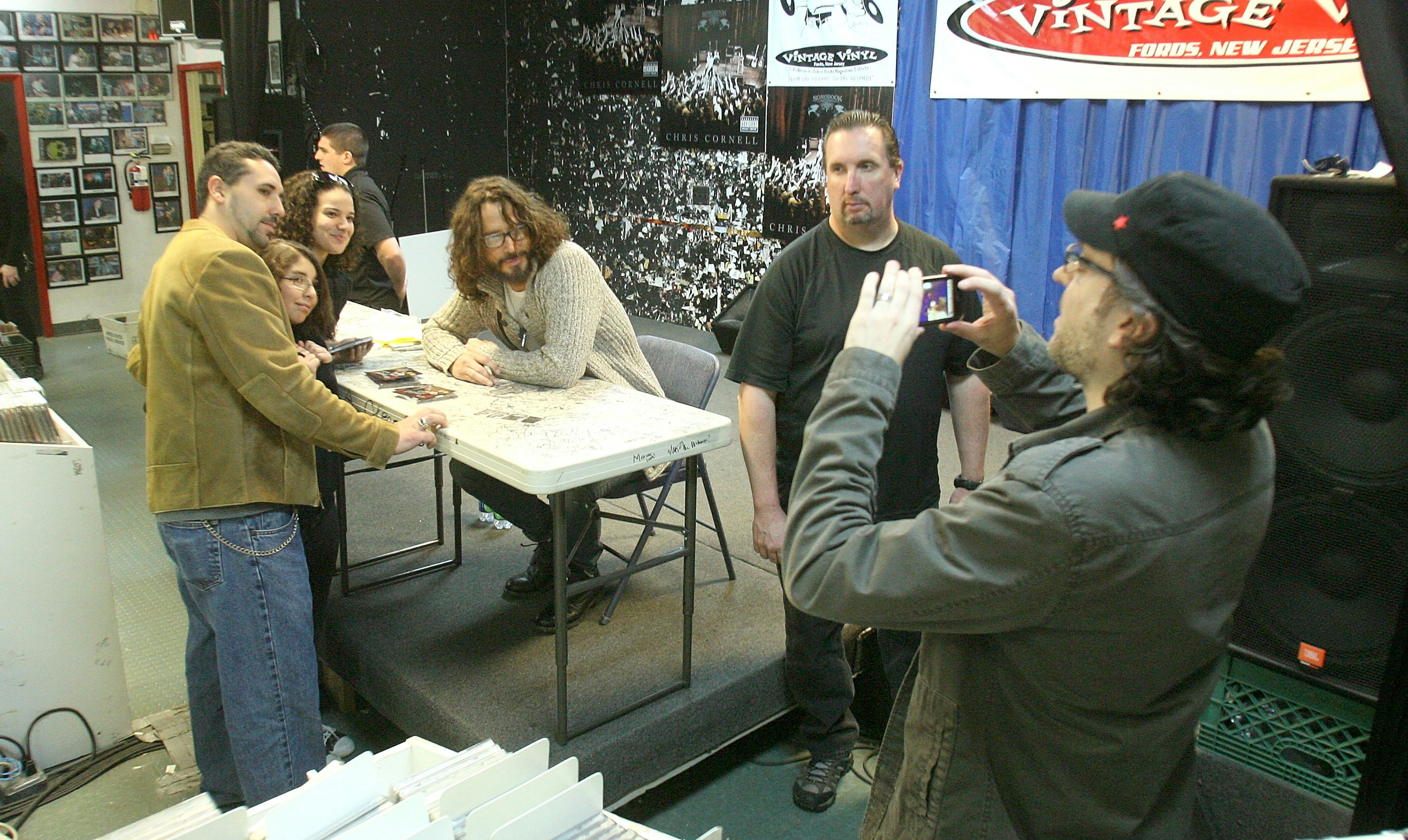 Musician Chris Cornell sits in an oatmeal colored sweater at a white card table in front of a blue backdrop and red and white Vintage Vinyl logo banner as a photographer in a black hat and grey shirt snaps a photo of Cornell with a fan in a tan jacket and jeans at a promotional event in 2011.