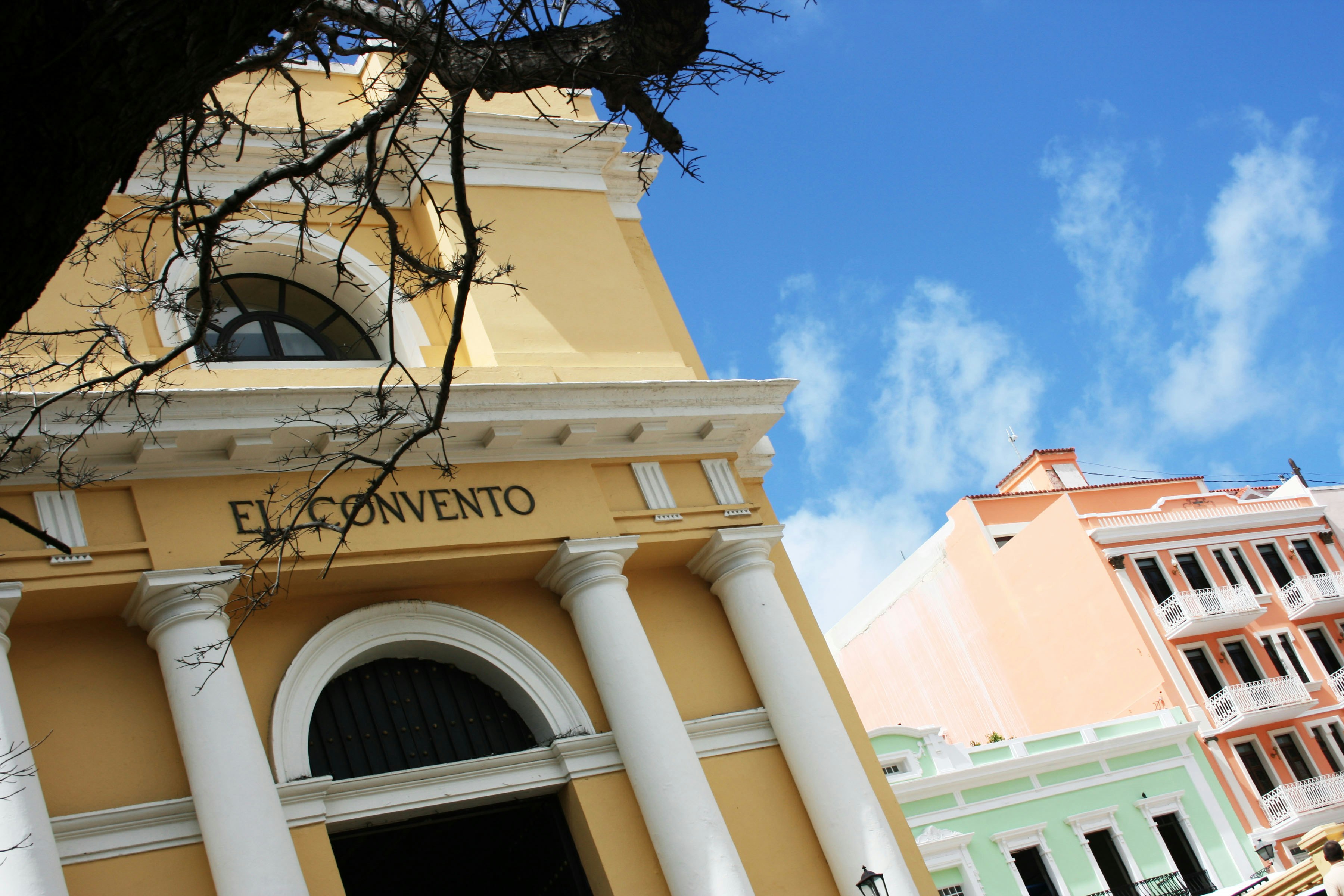 Close-up of a buttercream-colored neoclassical building, on which are the letters spelling out 'El Convento'. Other pastel-colored buildings can be seen behind it, beneath a blue sky