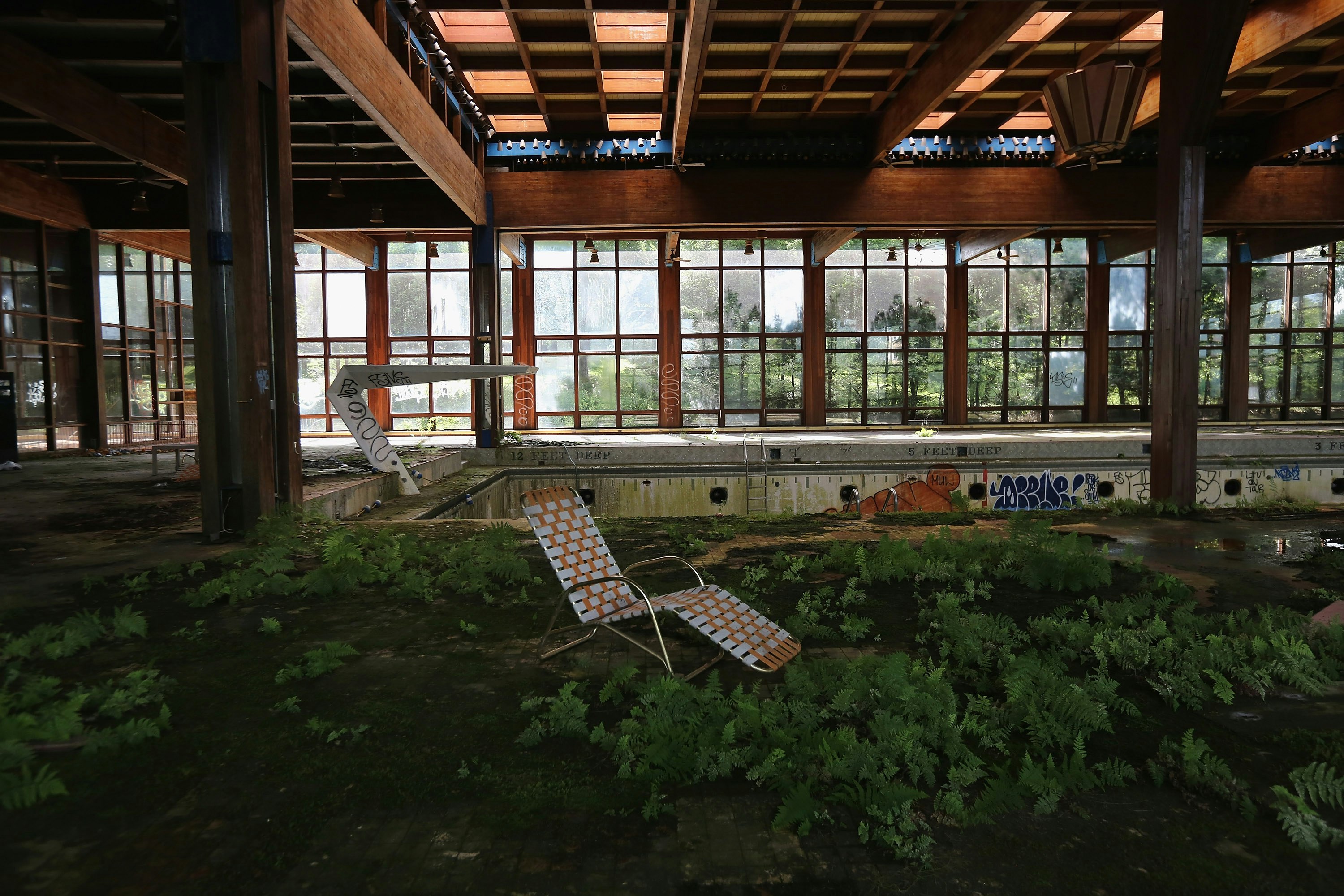 The once-luxe pool at Grossinger's resort is grown over, with ferns and ivy covering what was once the pool deck and graffiti dotting the drained concrete interior of the pool. A lone orange and white deck chair sits in the center of the frame hi-lighting the decrepitude.