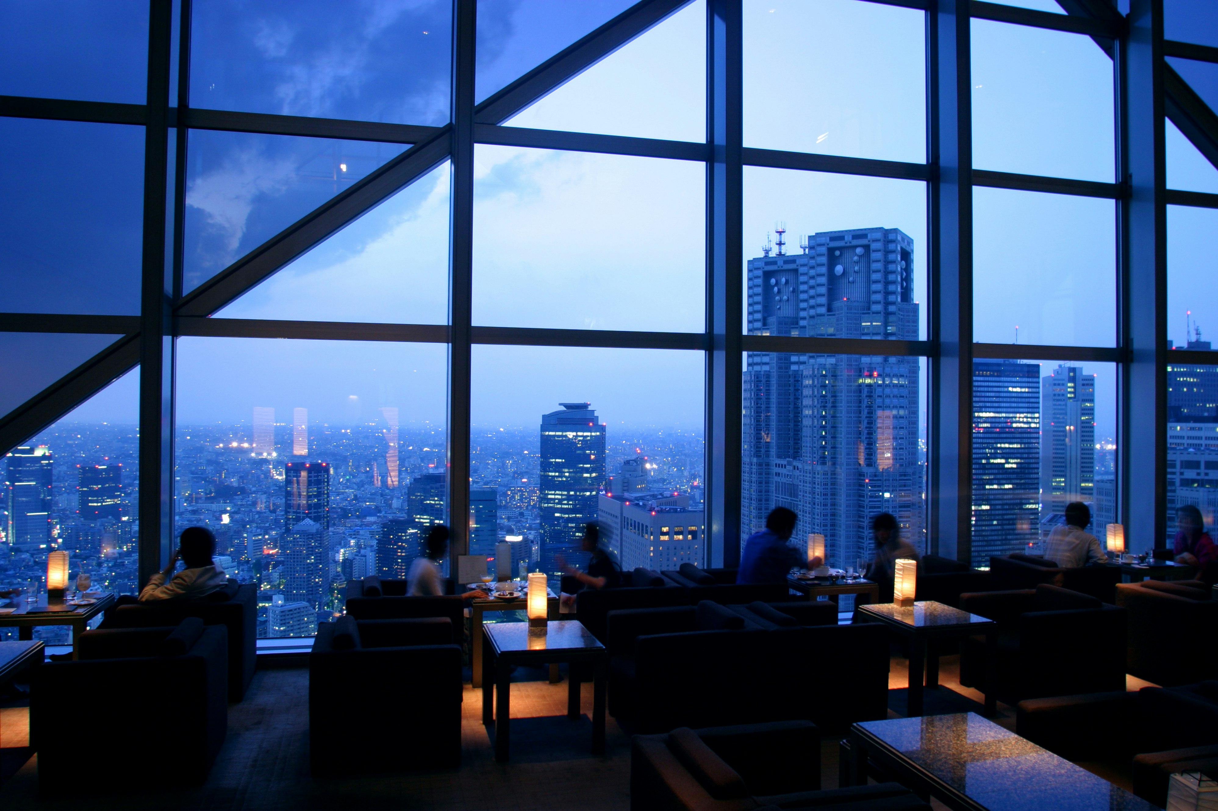 The skyline of Kanto district in Tokyo is bathed in twilight denim blues punctuated by white and red lights from the skyscrapers in view. A wall of windows divided into squares and triangles by large steel girders separates a few diners at small tables with easy chairs and rectangular lanterns from the view outside.