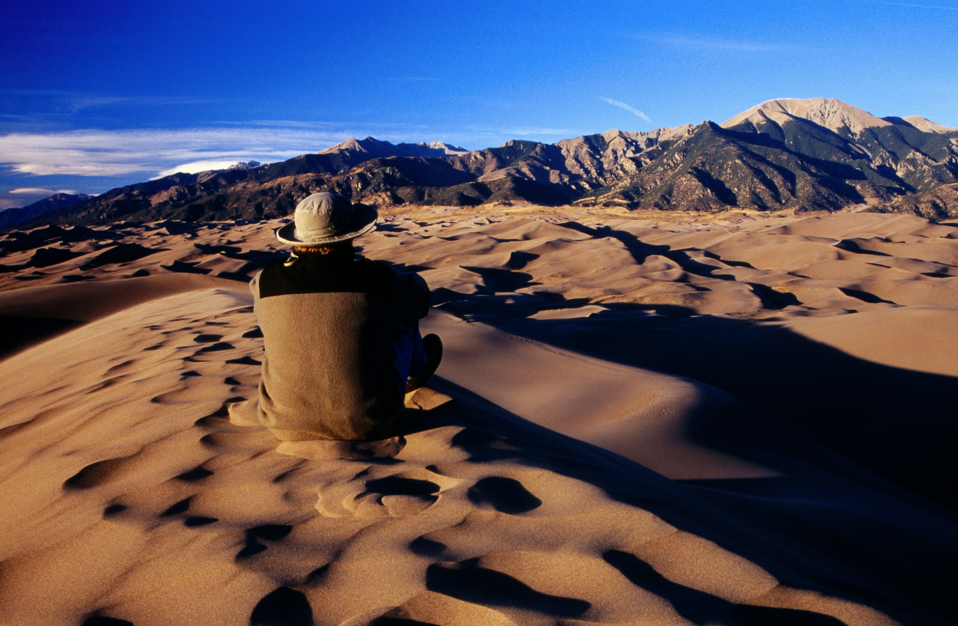 A man in a beige fleece jacket and a twill bucket hat sits with his back to the camera, blending into the sand dune on which he is perched, staring at the vast Sangre de Cristo Mountains in the distance.