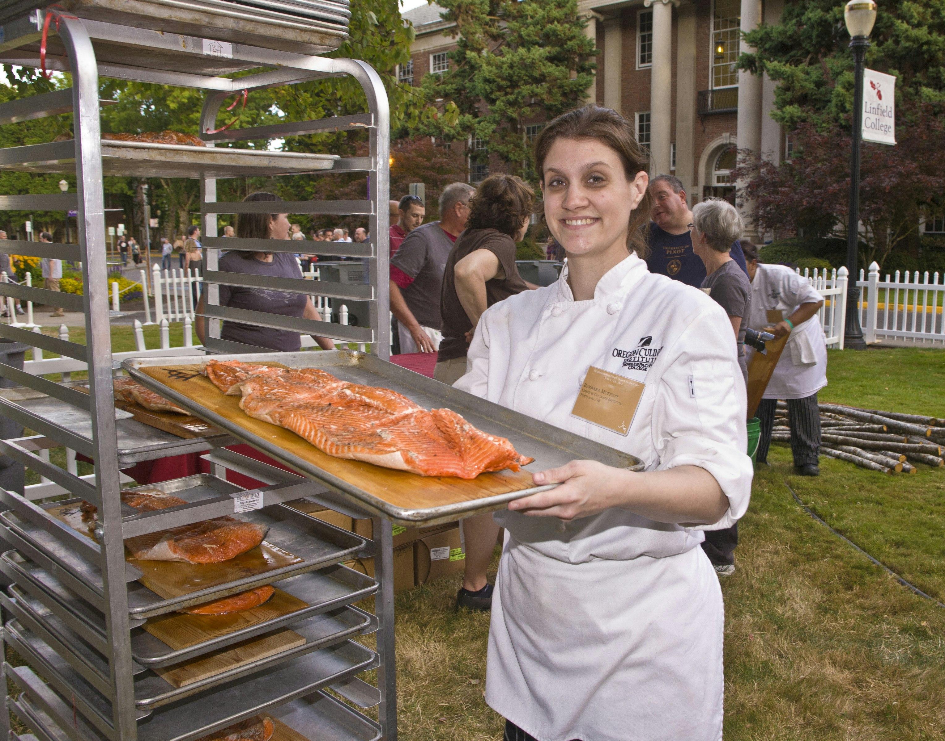 A woman if a white chefs coat pulls a tray of wood-fired salmon from an industrial cooking rack at the 2012 Pinot Noir Conference in McMinnville, Oregon 