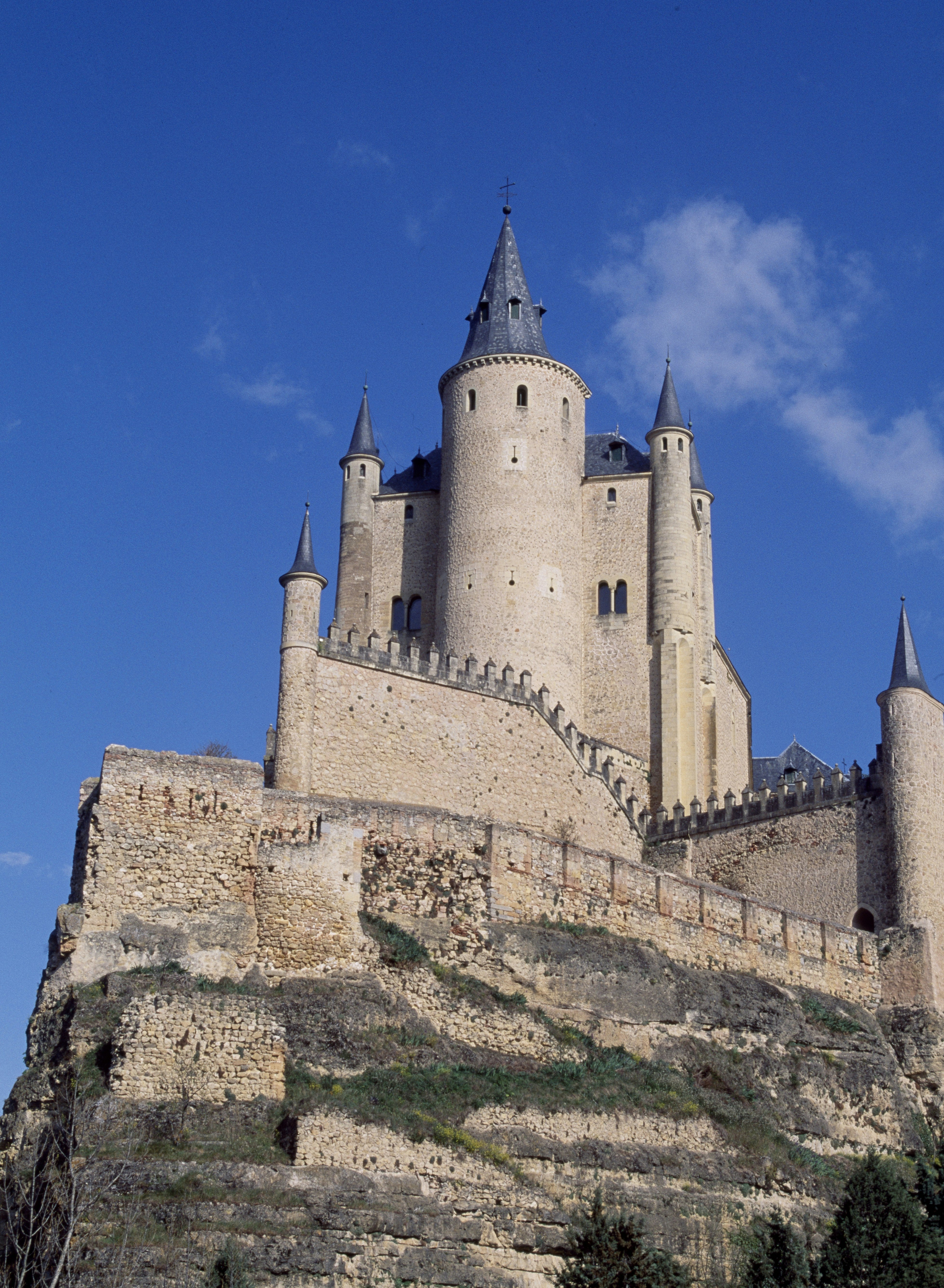 Alcazar de Segovia, a castle with dramatic, pointed spires, crenellated fortified walls, and rounded lookout towers is perched precariously on a rocky outcrop, beneath a blue sky with few clouds.