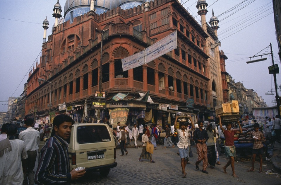 A man stands in front of the Nakhoda Masjid mosque on Rabindra Sarani street in Kolkata. The facade of the mosque is red clay or stone with scalloped edges around numerous columns and minarets. The roof is made up of pale blueish onion domes. Behind the man is a brown van, and banners in illegible Hinid hang from telephone wires over the intersection.