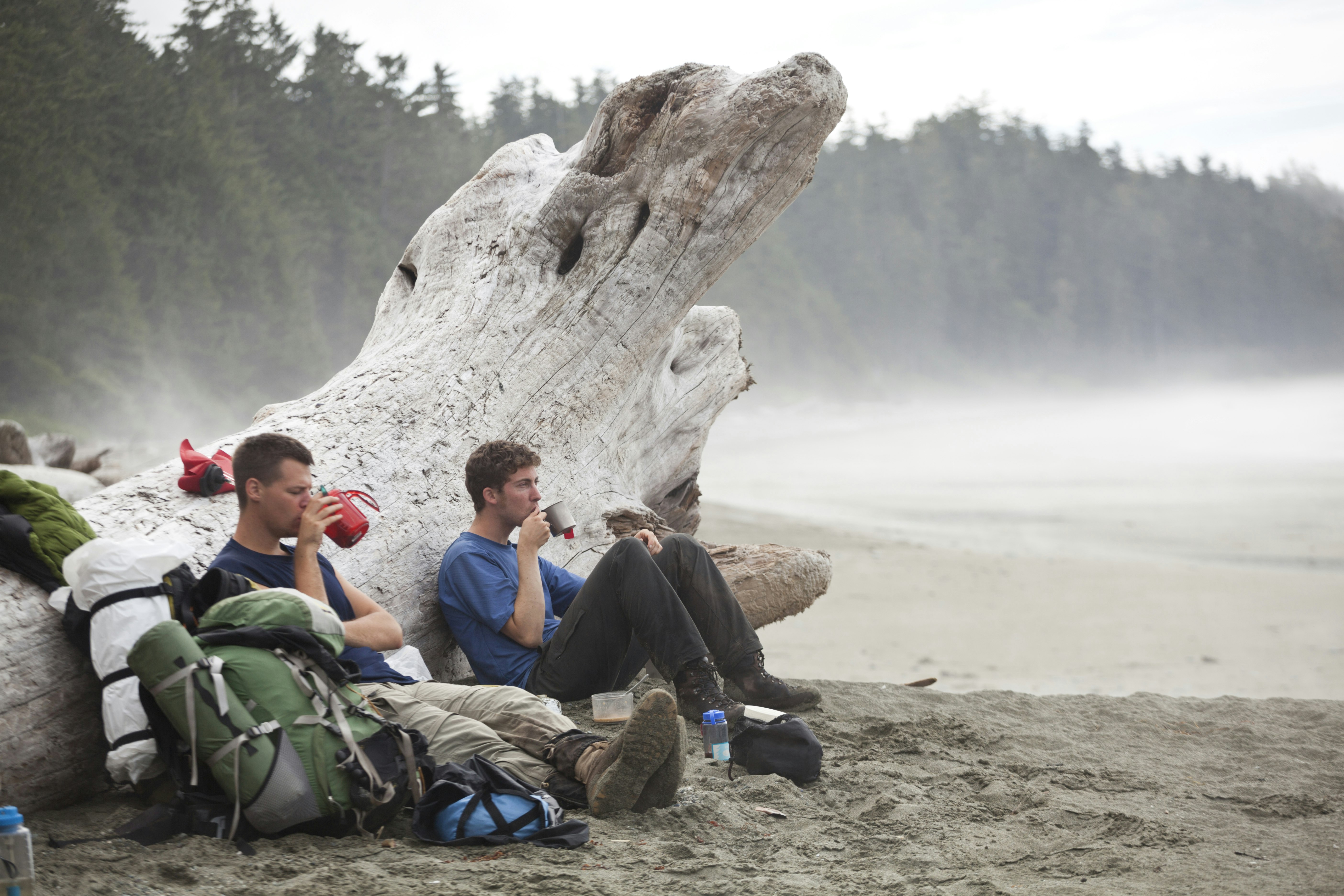Hikers rest on the West Coast Trail in British Columbia, sitting on the sand against a giant piece of driftwood as they sip from their mugs and gaze out at the foggy Pacific Ocean