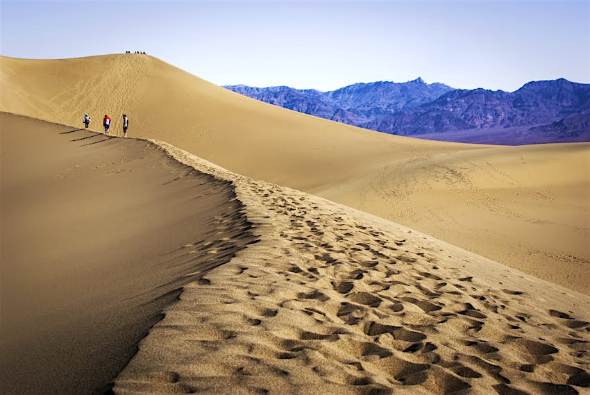 mesquite-dunes-death-valley-national-park Three hikers walking along a ridge amidst huge sand dunes. Another group of hikers is visible atop another dune in the distance while, beyond, is a craggy mountain range.