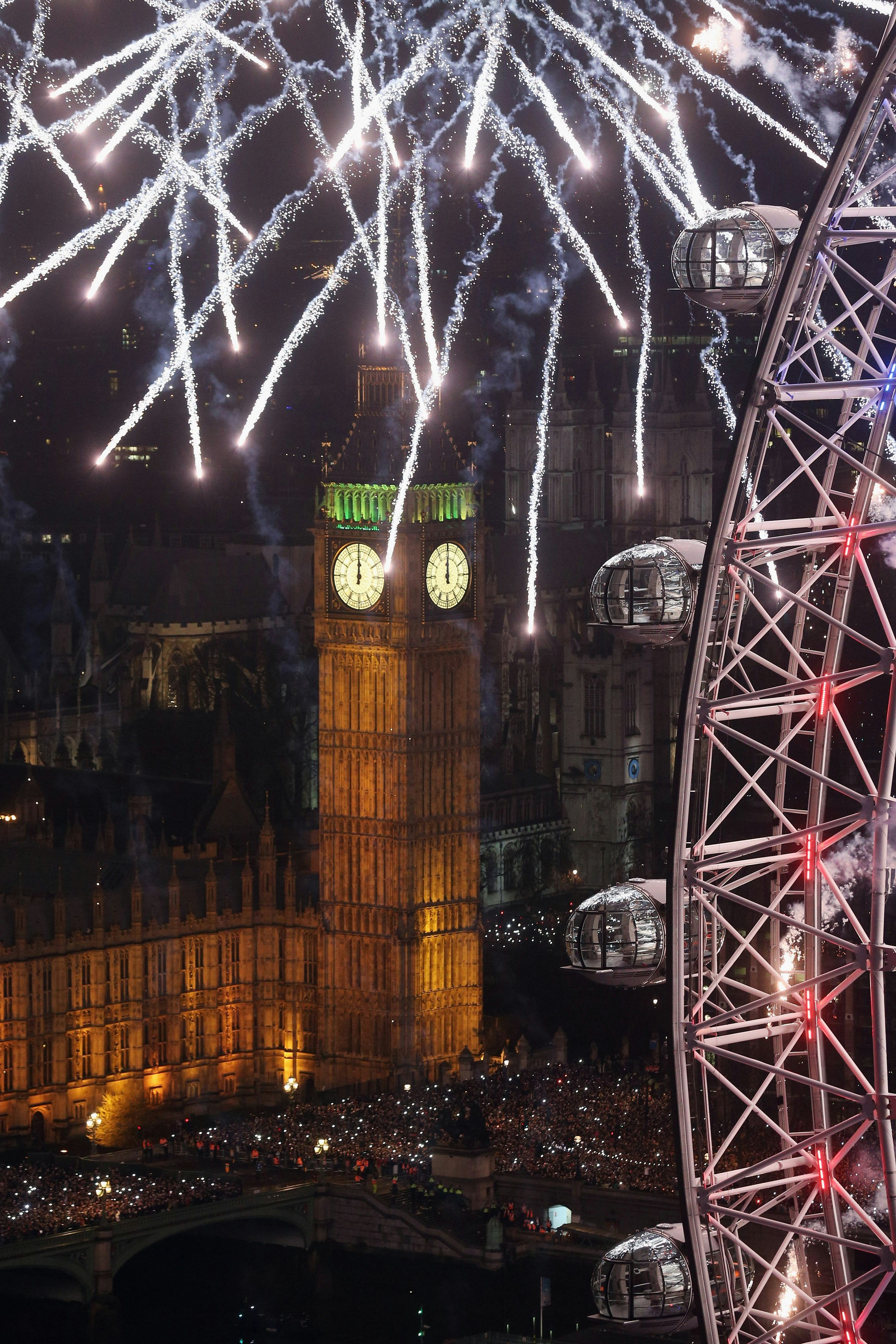 Aerial view of the New Year's Eve street party in London beside the Palace of Westminster. Part of the London Eye can be seen in the near foreground, while fireworks rain down over Elizabeth Tower (Big Ben). Thousands of people are gathered in the streets below.
