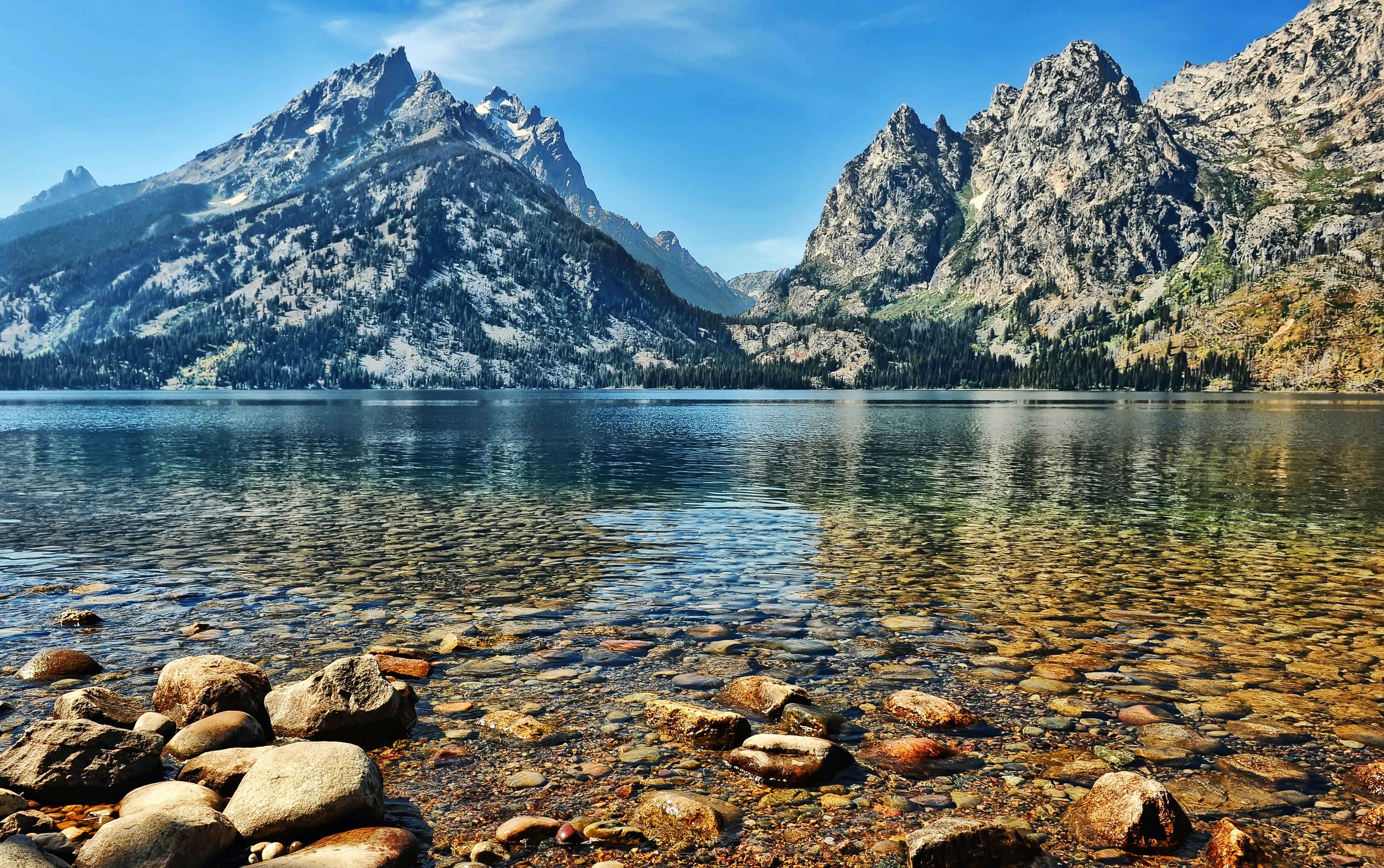 The ragged peaks of Grand Teton National Park rise steeply over Jenny Lake. The massif on the left is deep blue in the shadows and where trees cover its flanks, while the bright sunshine hi-lights its light grey rocky face. On the right, another peak is more brightly lit, appearing a light tan color with thin streaks of bright green vegetation. The lake itself is smooth and calm, a mixture of blues and greens, while in the extreme foreground the large, flat, stones on the shoreline are rust colored and grey