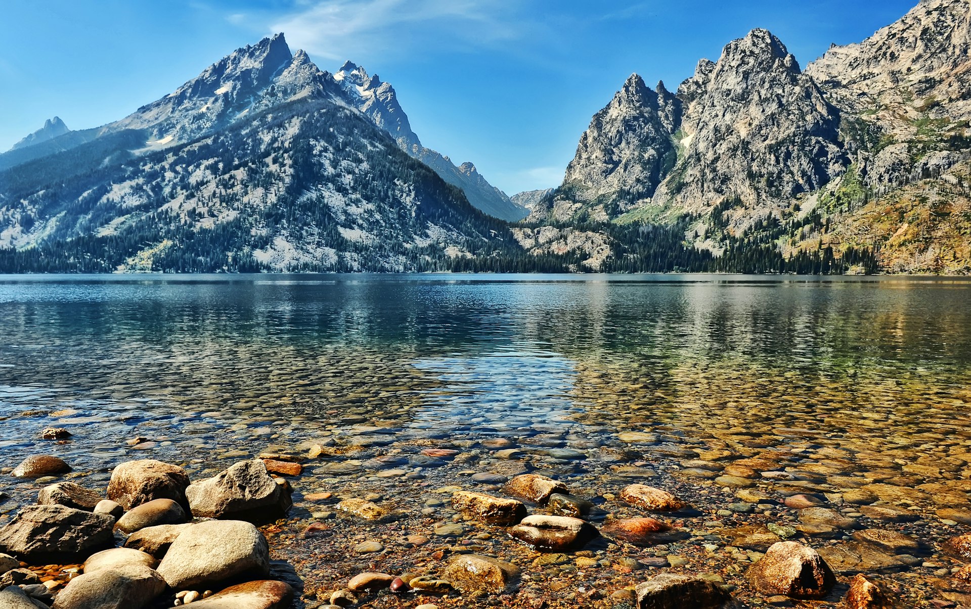 The ragged peaks of Grand Teton National Park rise steeply over Jenny Lake. The massif on the left is deep blue in the shadows and where trees cover its flanks, while the bright sunshine hi-lights its light grey rocky face. On the right, another peak is more brightly lit, appearing a light tan color with thin streaks of bright green vegetation. The lake itself is smooth and calm, a mixture of blues and greens, while in the extreme foreground the large, flat, stones on the shoreline are rust colored and grey