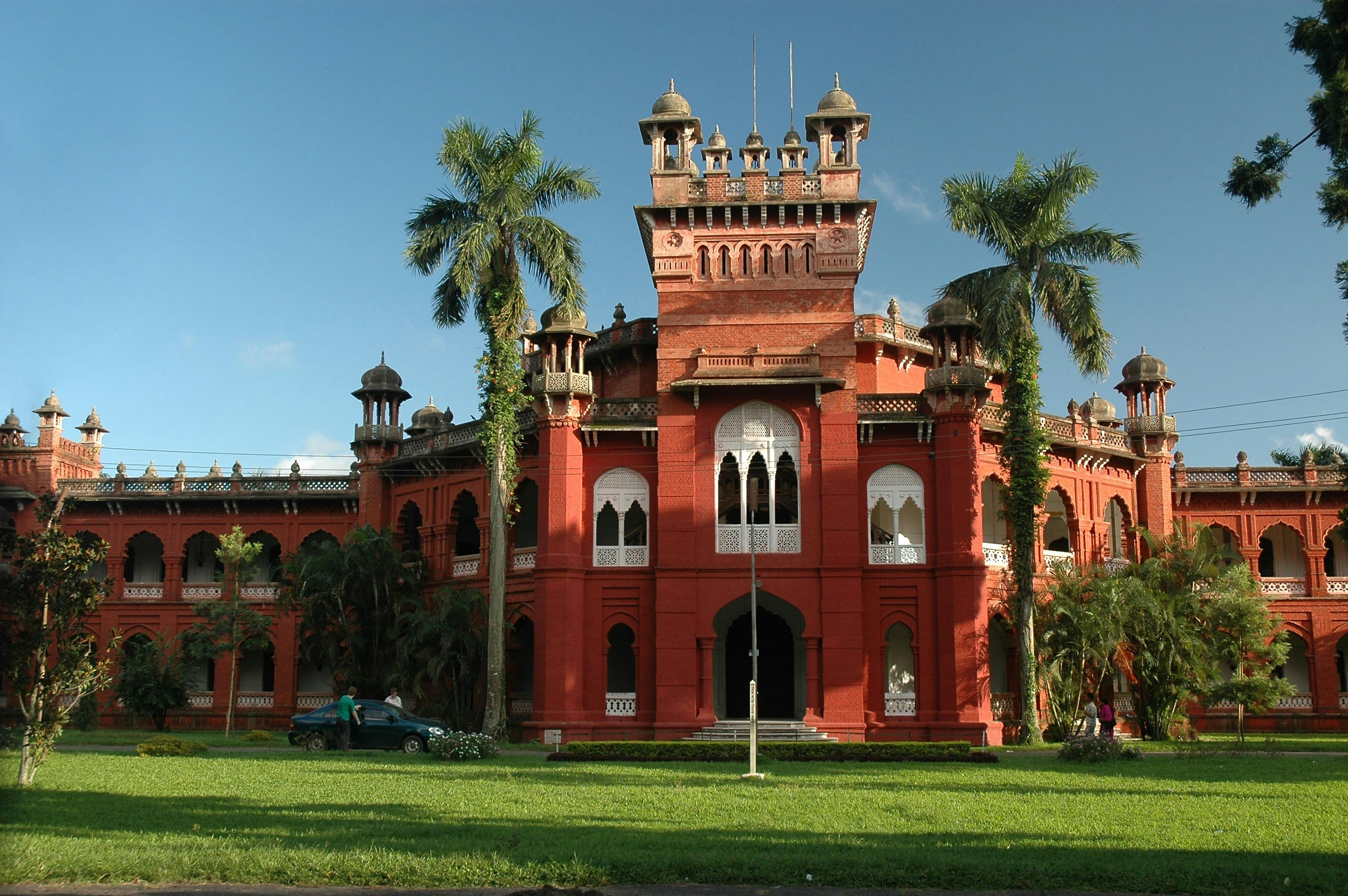The brick-red Curzon Hall, a quasi-Gothic building built in the style of European-Mughal architecture. The large building almost resembles a castle, with turrets, arched balconies and minarets. In front of the grand building is a manicured green lawn and some trees.