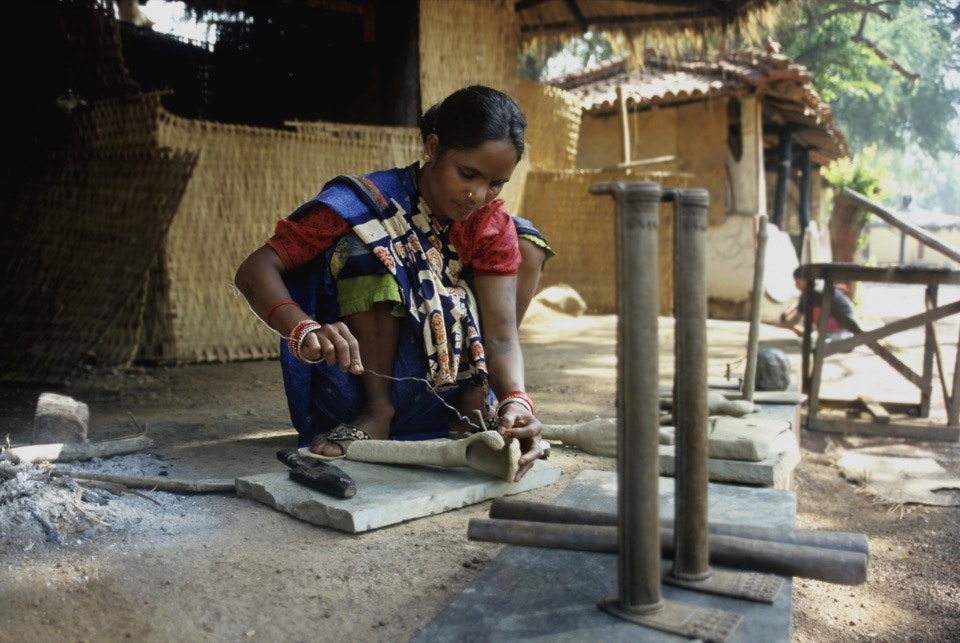 A woman in a colorful sari squats over a rough metal cast of an arm set on a stone slab, and is applying a metal wire to it as she works on her sculpture in front of woven reed mats.
