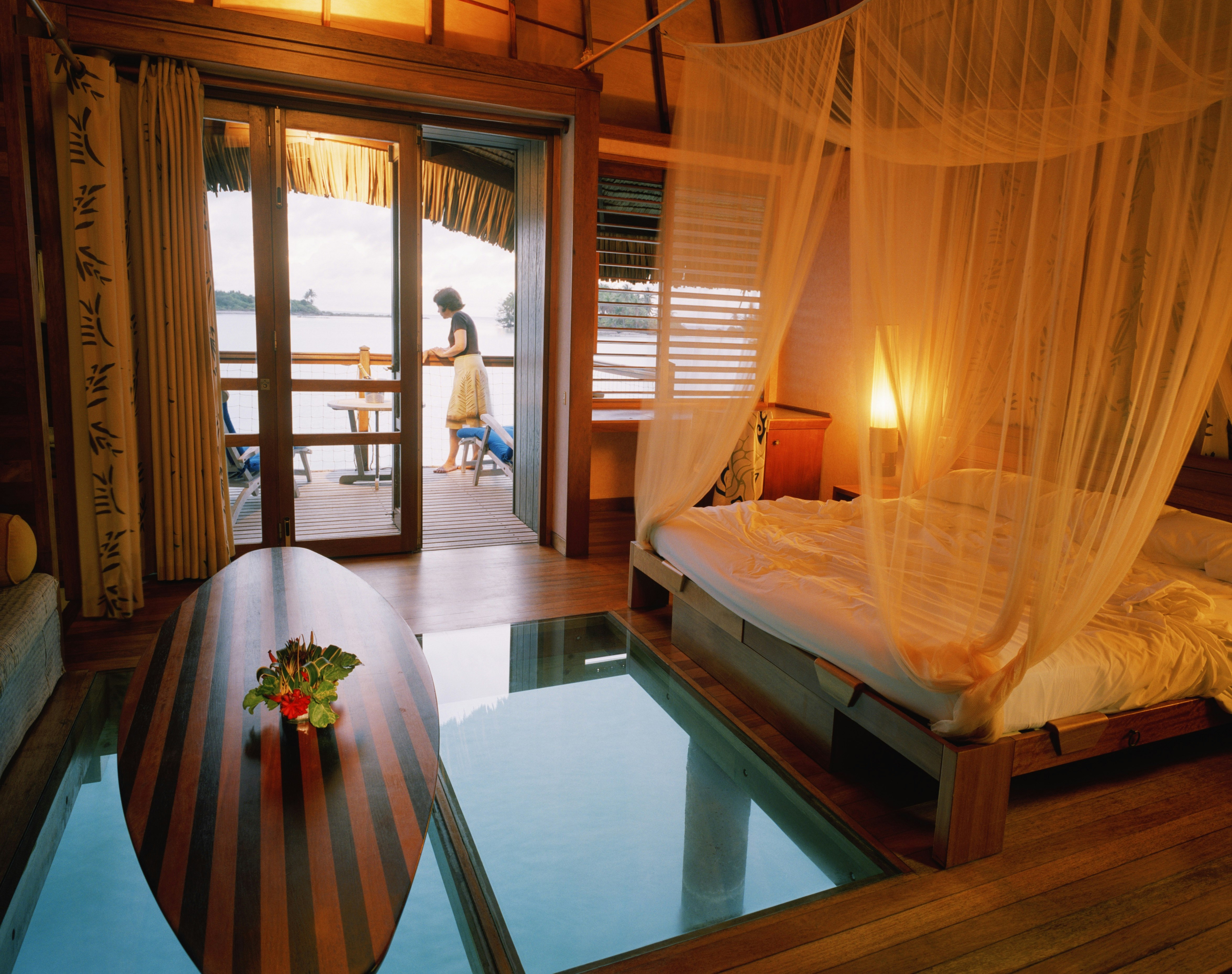 An overwater bungalow in Hotel Le Meridien in Bora Bora, where there are not only tropical views out the windows and off-deck, but also underwater via a glass floor. The bed is a simple wood platform with white sheets and mosquito netting. The wood is wide planks in a similar warm tone. A surf-board shaped coffee table of inlaid light and dark wood sits over the glass floor window. On the deck, a woman in a white skirt and black top pauses by the railing.
