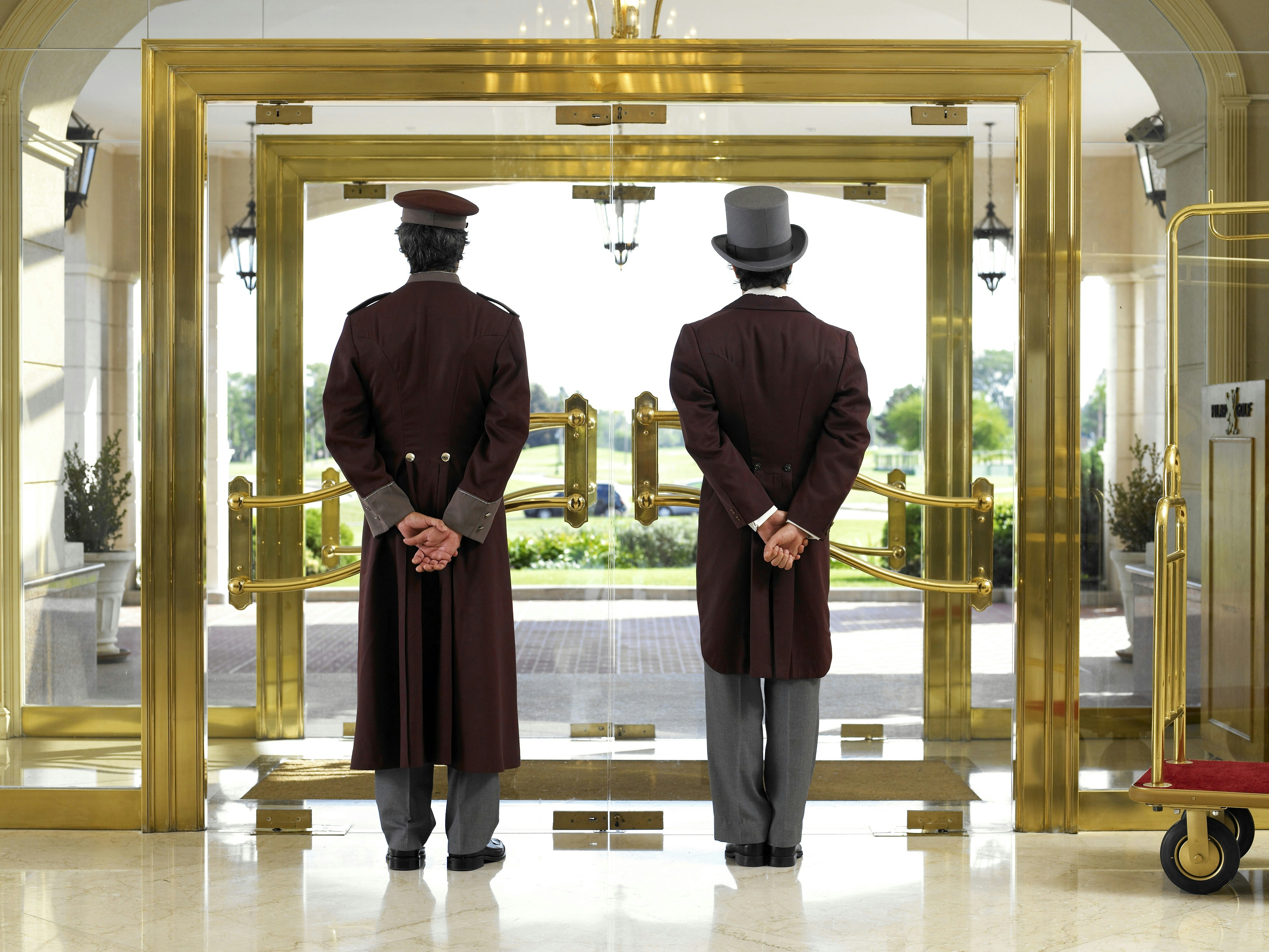A concierge and bellboy wait at the entrance to a hotel. Both are dressed in brown suits, with their backs to the camera. One wears a smart grey top hat. Through the glass doors some greenery is visible.
