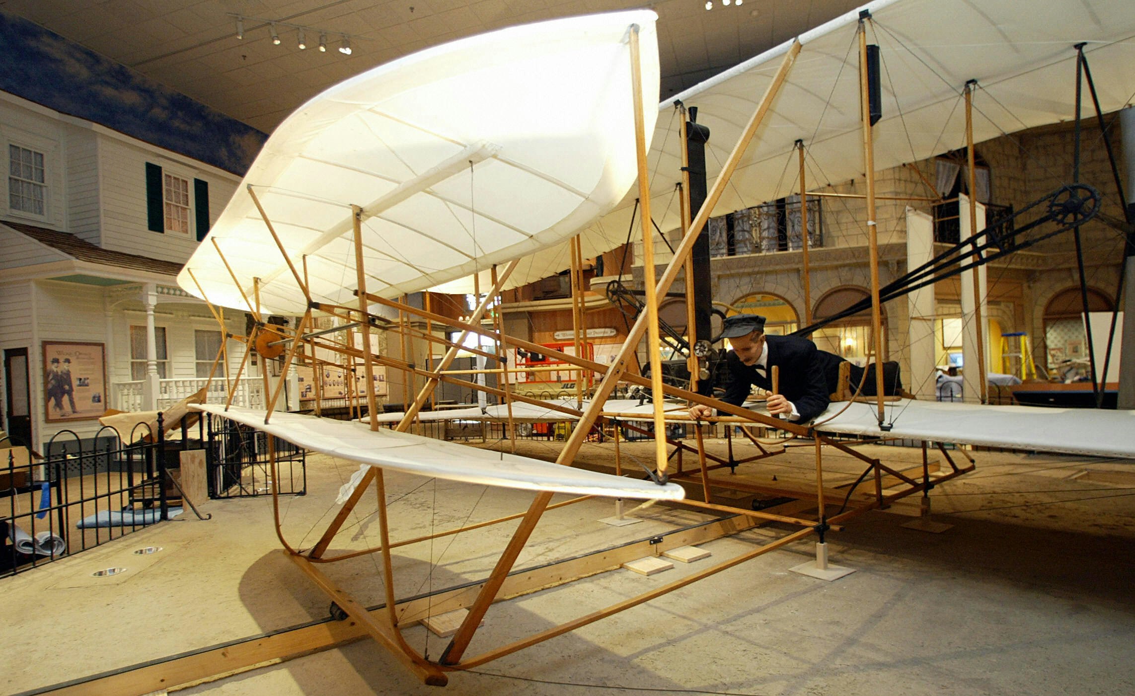 The 1903 Wright Flyer, the world's first airplane, on display at the Smithsonian's National Air and Space Museum in Washington, DC. It is made of light-colored wood slats with white material covering the wings. A mannequin lays belly-down on the plane as if about to take off. 