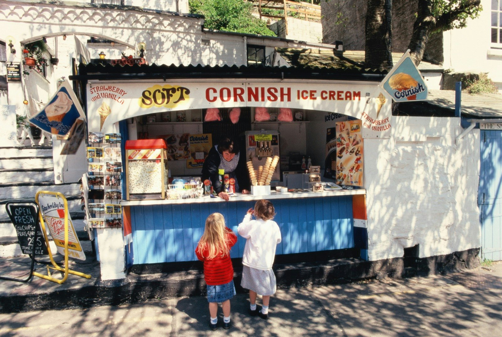 Two small children stand in front of a ice cream stand painted blue with red writing that says Cornish Ice Cream on a trip in Cornwall with toddlers