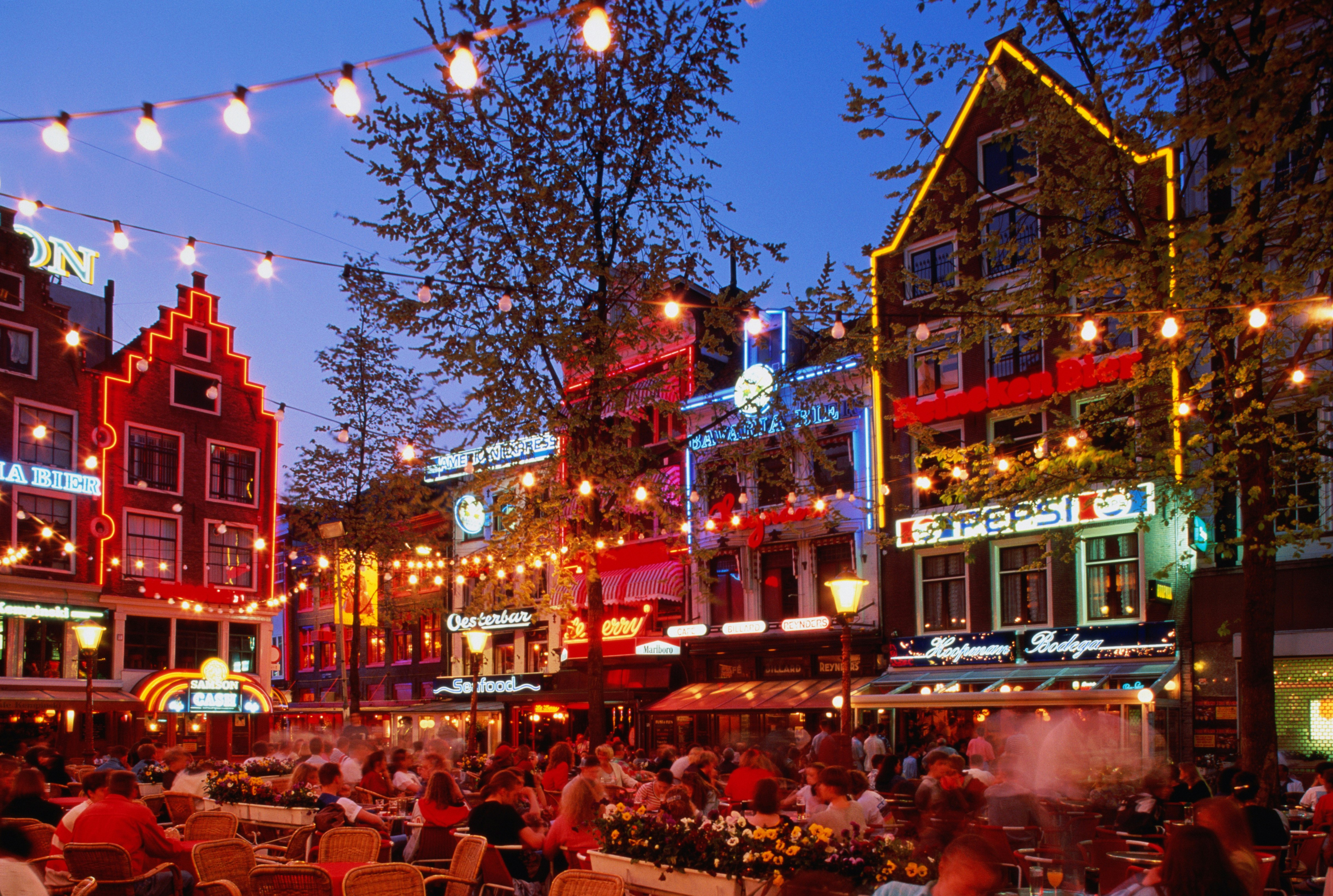 Ett fullsatt torg på Leidseplein på natten.  Bord och stolar, fyllda med människor, upptar torget medan sagoljus hänger ovanför och barer och restauranger kantar väggarna runt det.