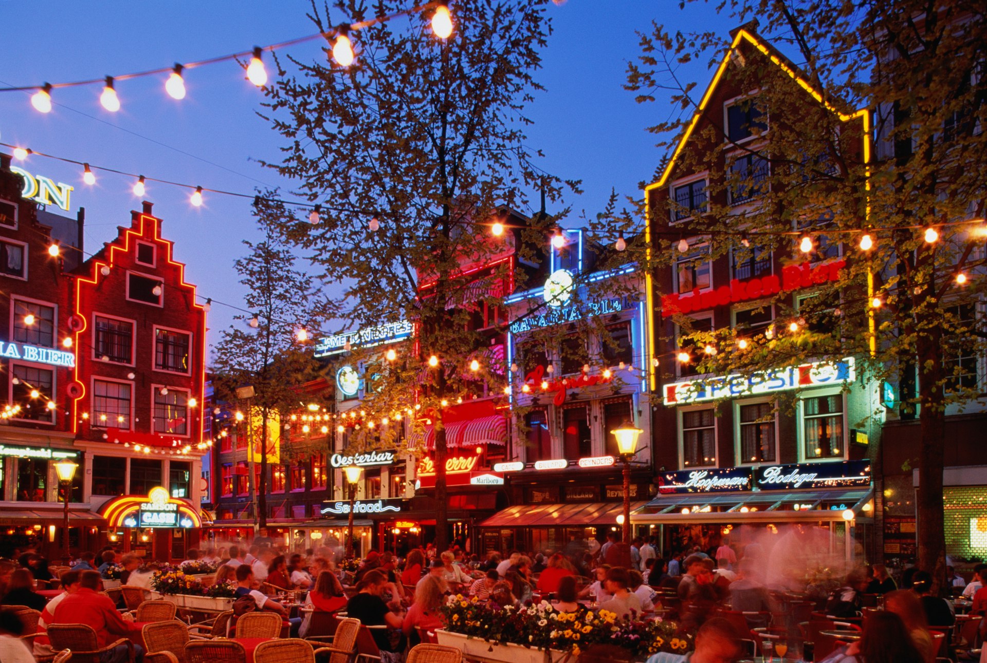 A crowded square in Leidseplein at nighttime. Tables and chairs, filled with people, occupy the square while fairy lights hang overhead and bars and restaurants line the walls around it.