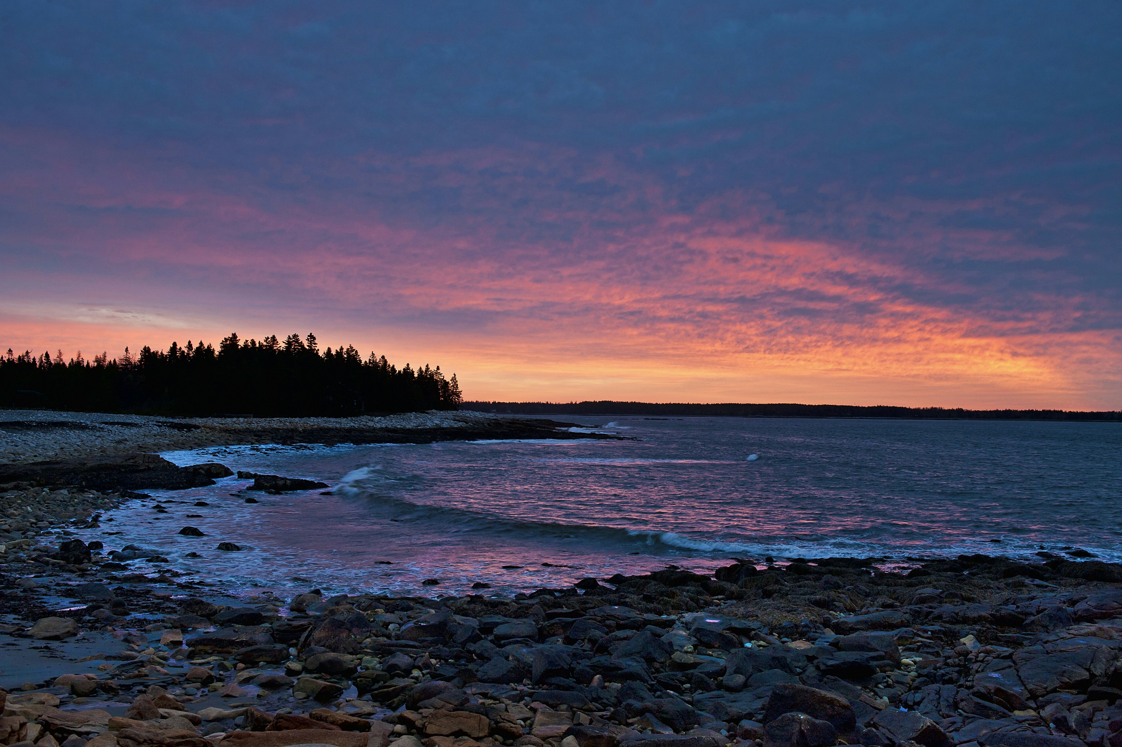 A brilliant sherbert pink and orange sunset reflects of dark blue clouds over Otter Bay in Acadia National Park in Maine. The ocean reflects the sunset in muted tones and a tall stand of trees is jet black in the background. Scuba diving in national parks