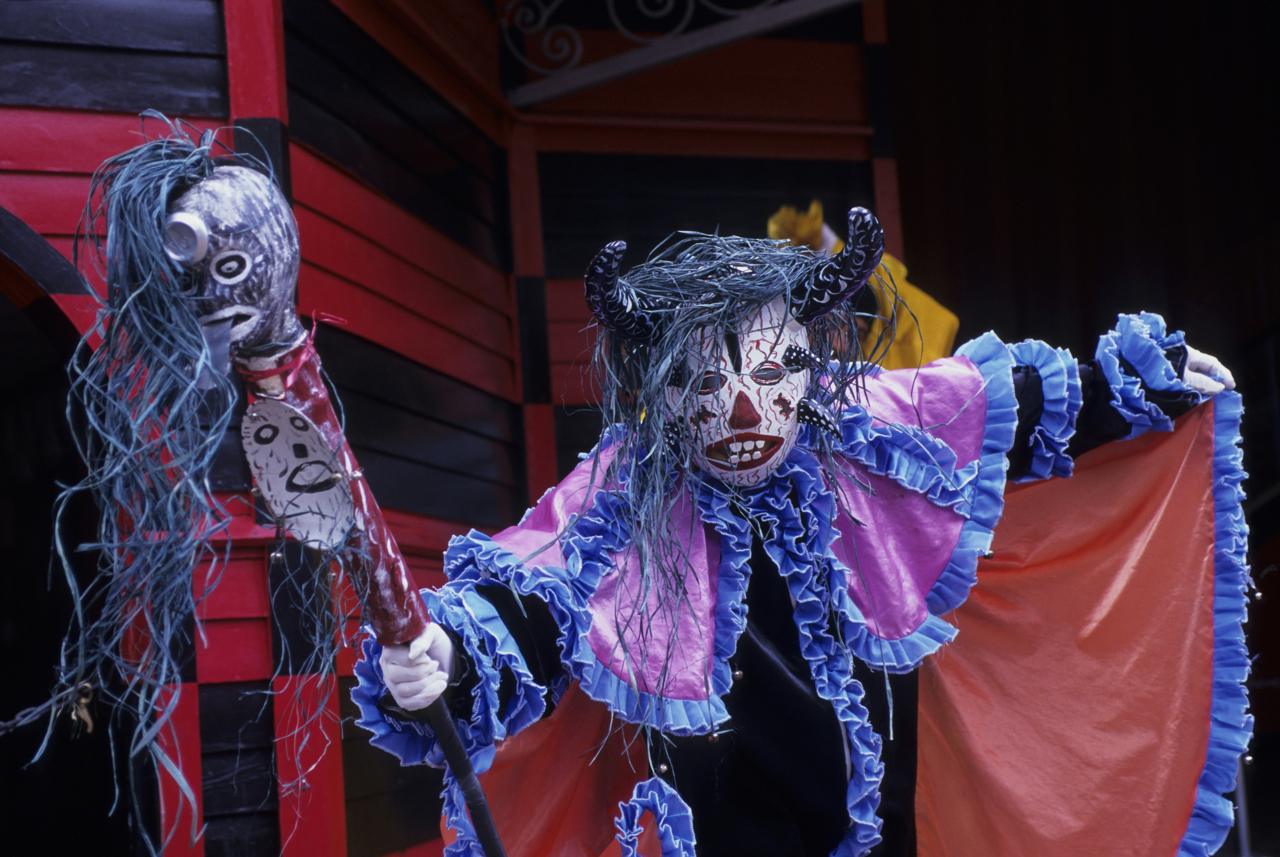A performer wearing a red and blue cap, wearing a horned face mask and carrying a staff leans menacingly towards the viewer, part of Afro-Puerto Rican festival custom