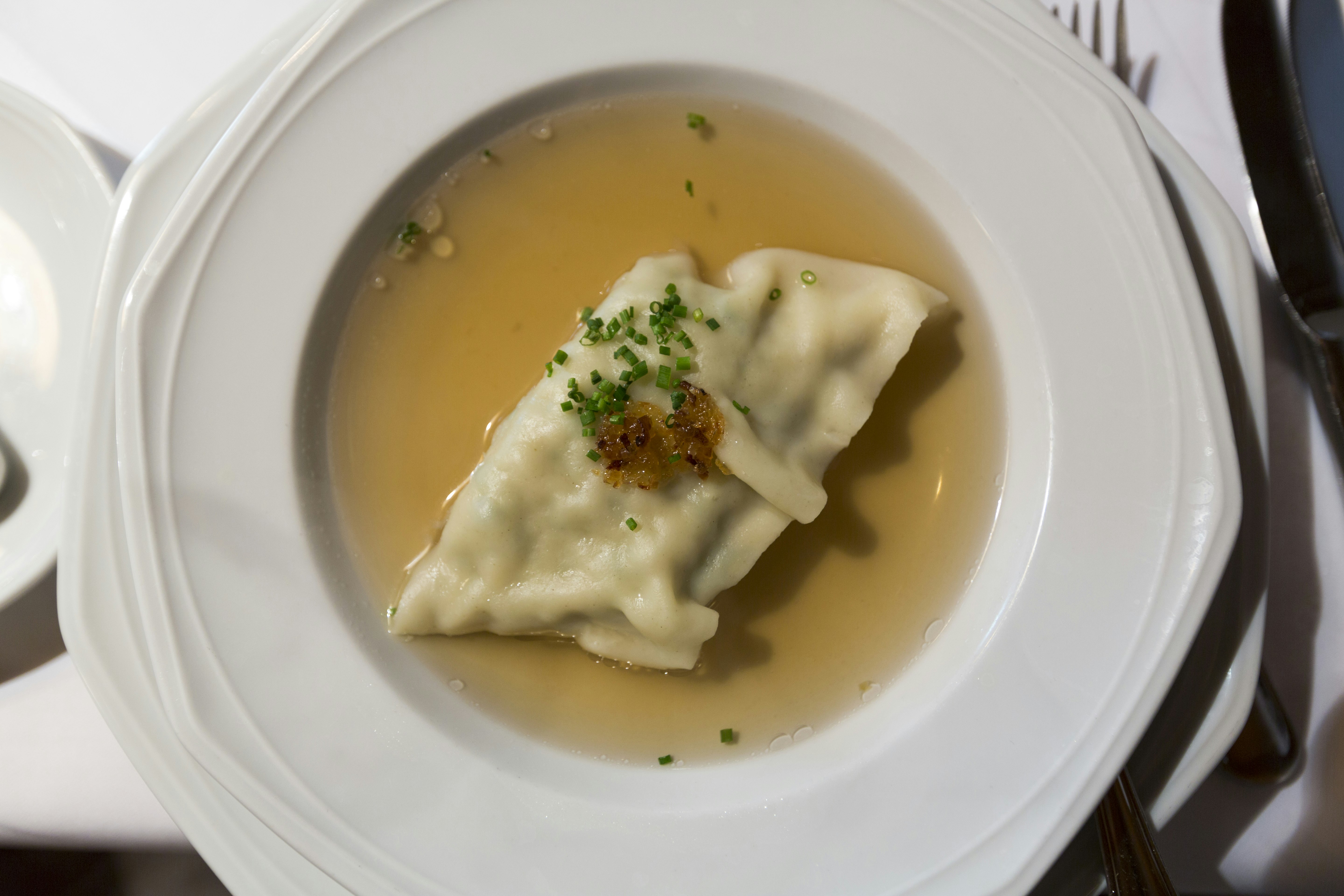 A pale dumpling topped with dark brown spices and bright green herbs sits in a pool of golden broth in a large white serving dish on a white table cloth.