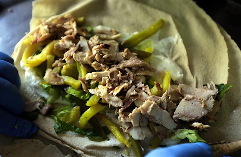 A close-up of someone wearing blue latex gloves, who is preparing to roll a chicken shawarma. Pita bread is loaded with jalapenos and shredded chicken.