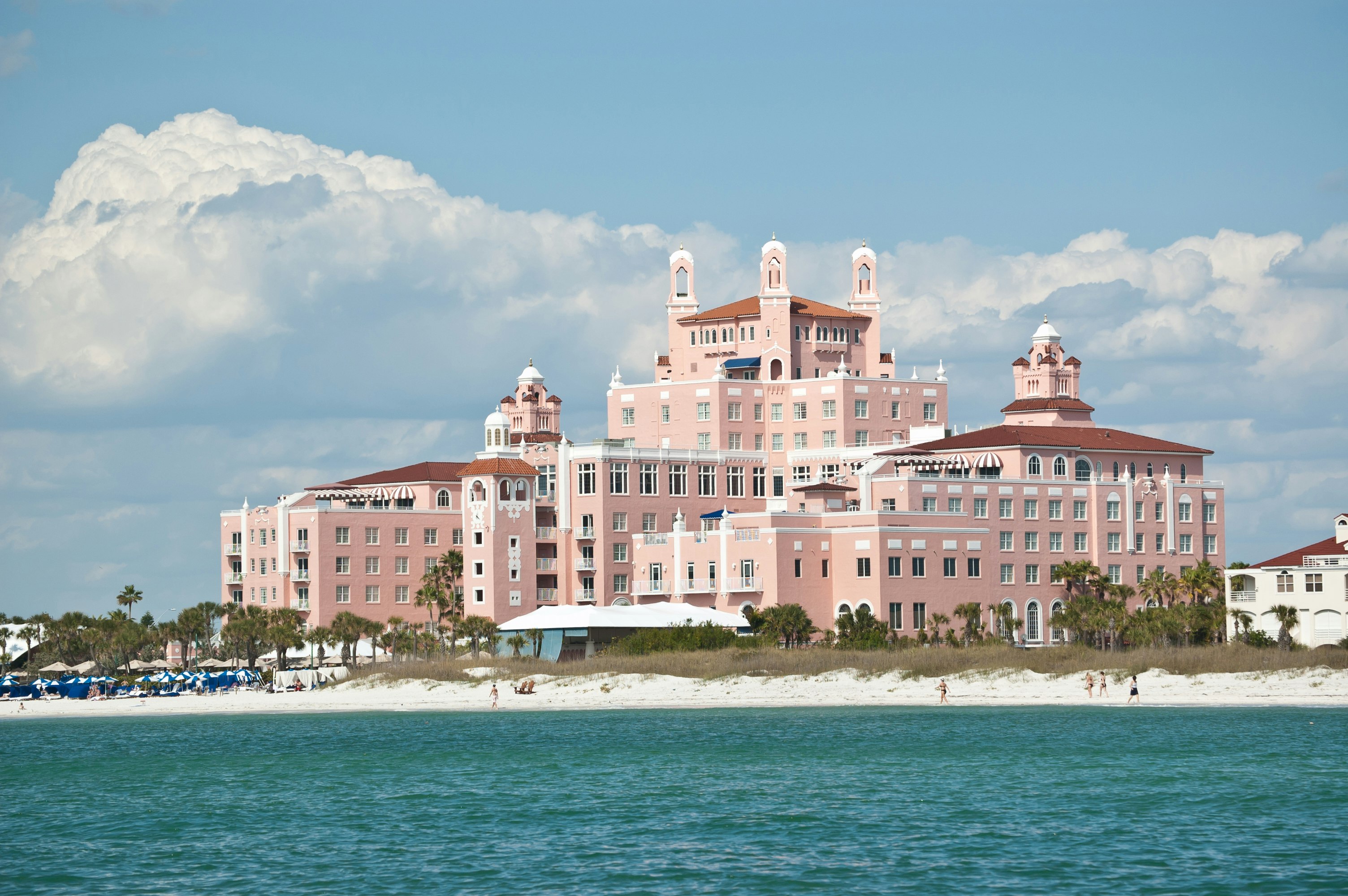 The pink art deco facade of the Don CeSar Resort rides over the white sands of St. Pete beach in Florida and the deep aquamarine waters of Tampa Bay, with big white cumulonimbus clouds against the stone-washed denim sky in the background.