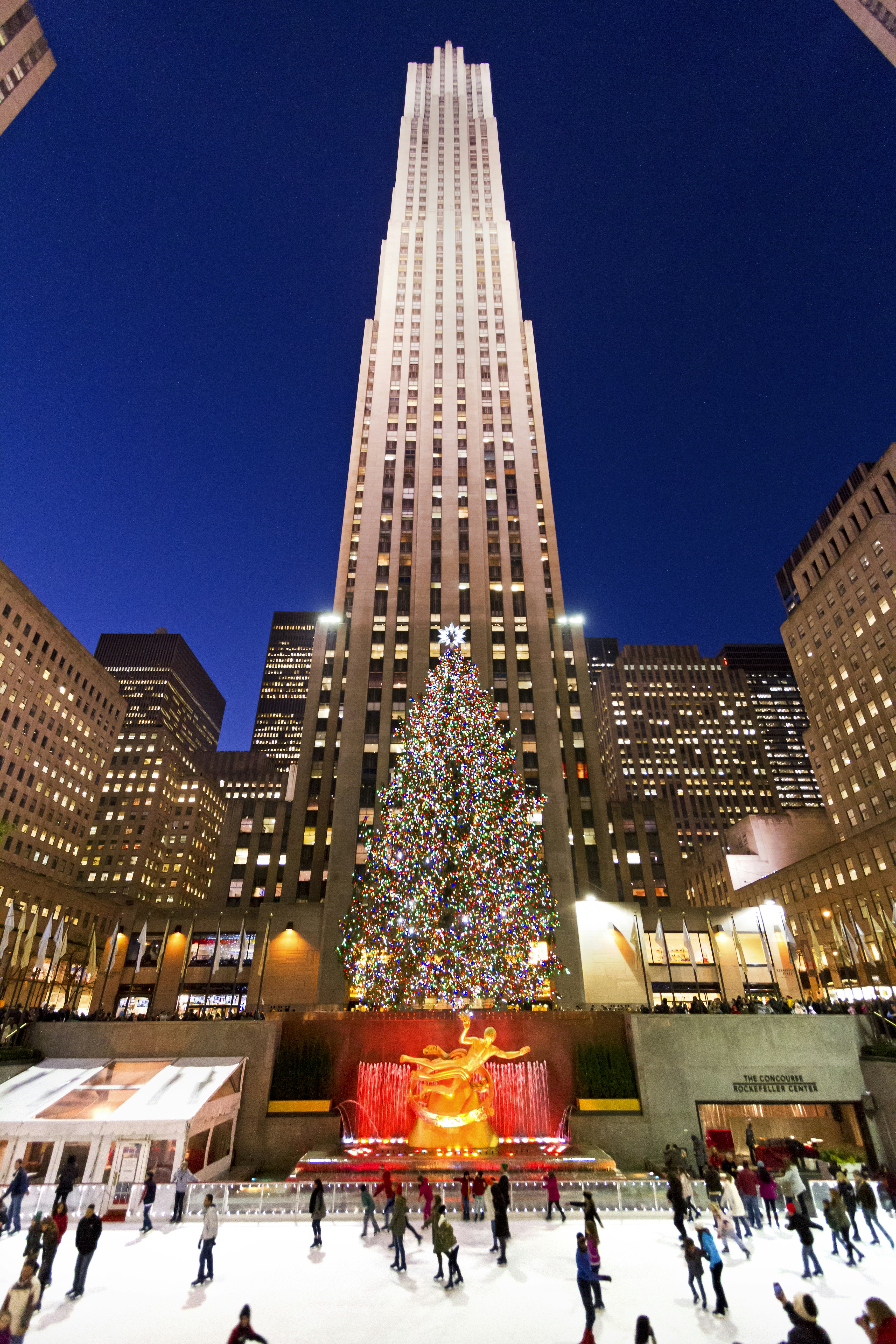 A tall building stands behind a large colorfully lit Christmas tree in front of which is the golden statue of Prometheus while people skate in front 