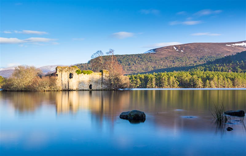 A view from the banks of Loch An Eilein, looking out towards the ruined castle on an island in the middle of the Loch. Beyond, the opposite forest-lined bank is visible, leading away to rolling mountains.
