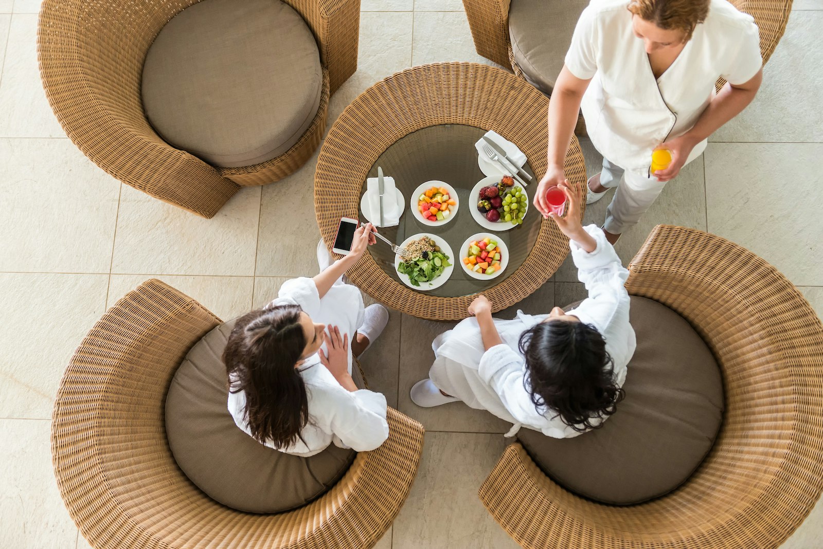 Two girlfriends in white bathrobes and slippers eating a healthy spa lunch at a resort hotel while a therapist serves them fresh orange and watermelon juice. Aerial, overhead view.