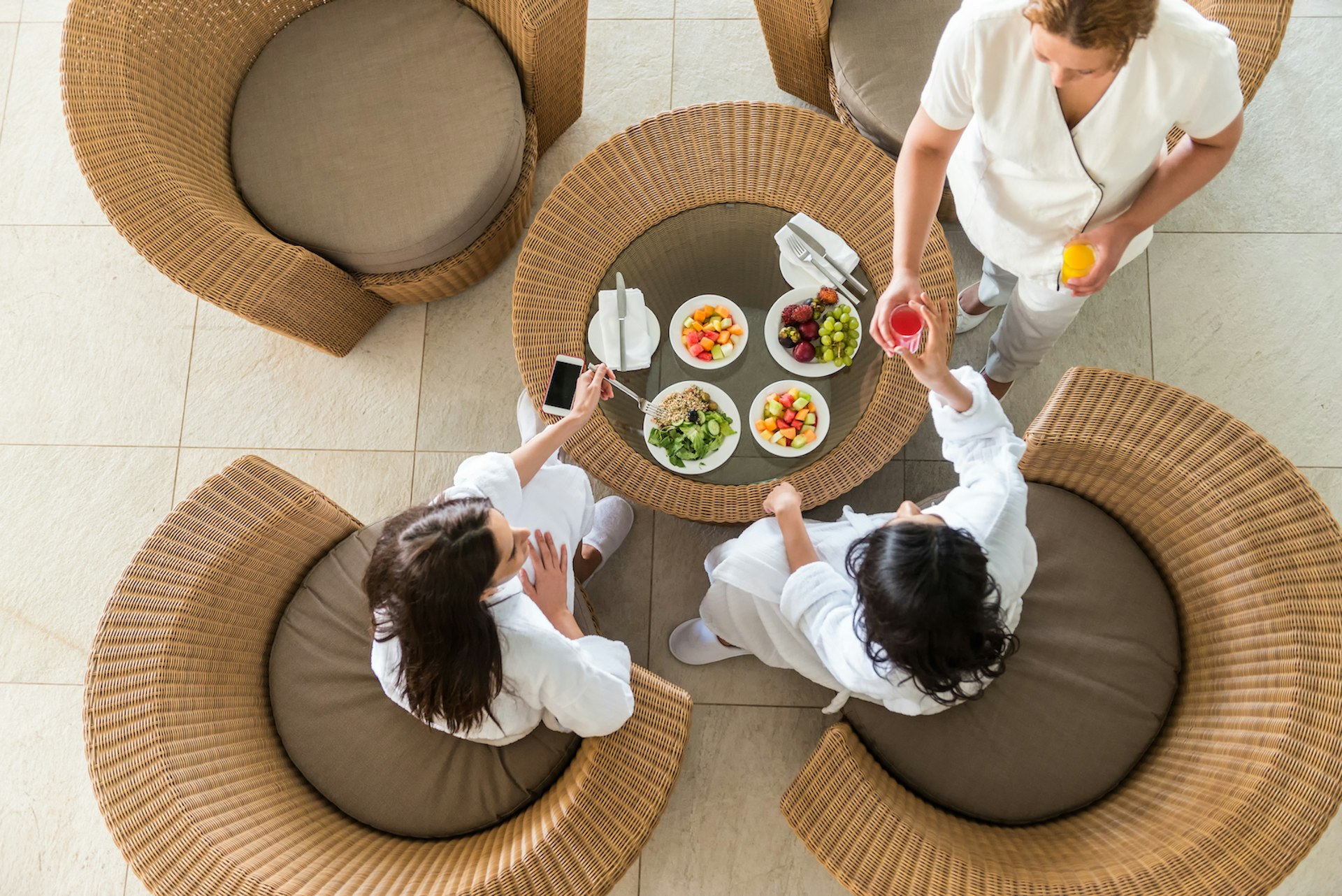Two girlfriends in white bathrobes and slippers eating a healthy spa lunch at a resort hotel while a therapist serves them fresh orange and watermelon juice. Aerial, overhead view.