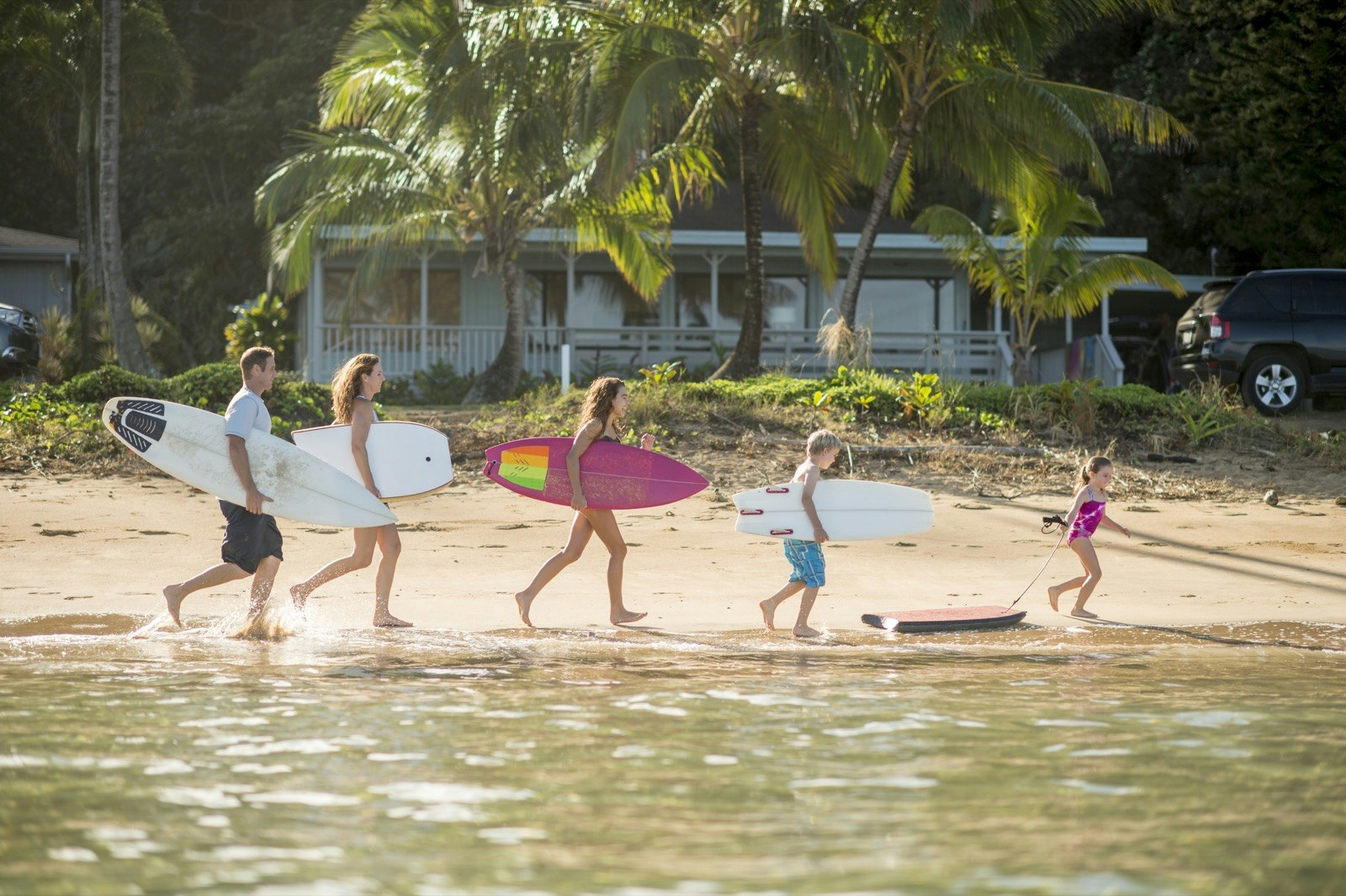 a family of five runs down a beach with a cottage in the background each one holding a surfboard or boogie board