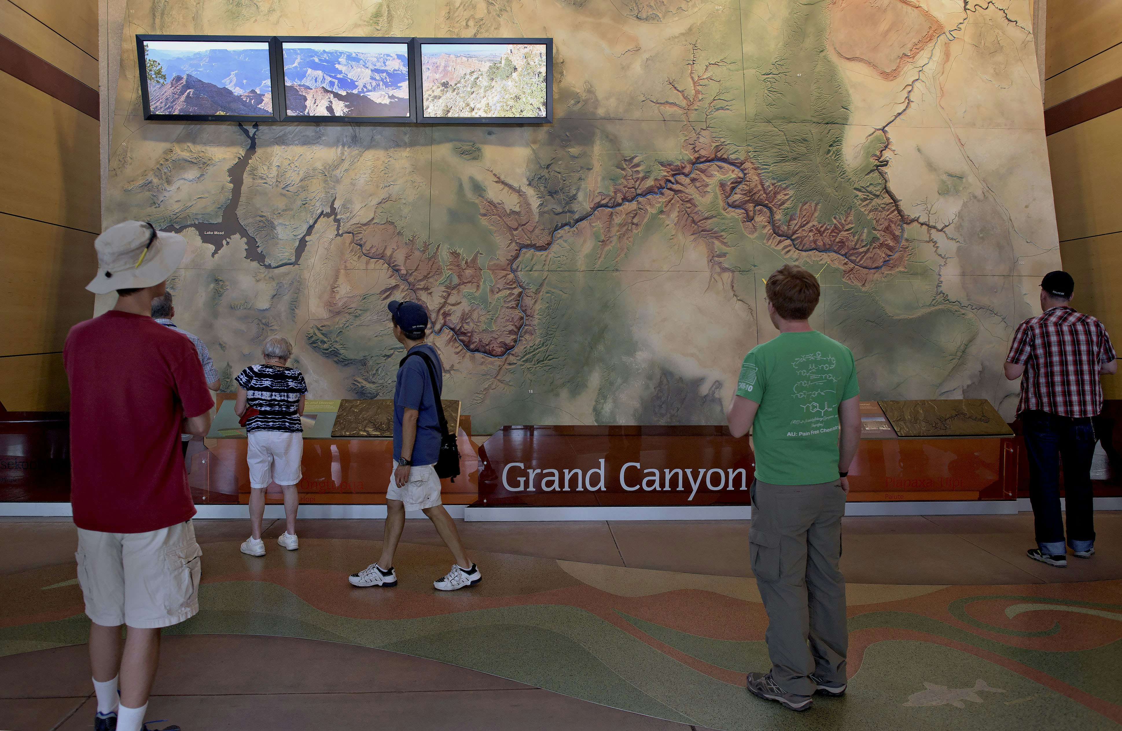 Six tourists stand in front of a large, wall-sized topographic map of the Grand Canyon with three television screens in its upper left corner to show footage of the park. On the floor, an abstract design in the same earth-tone shades of green, sage, orange-red, and buff as the map swirl in stone composite on the floor.
