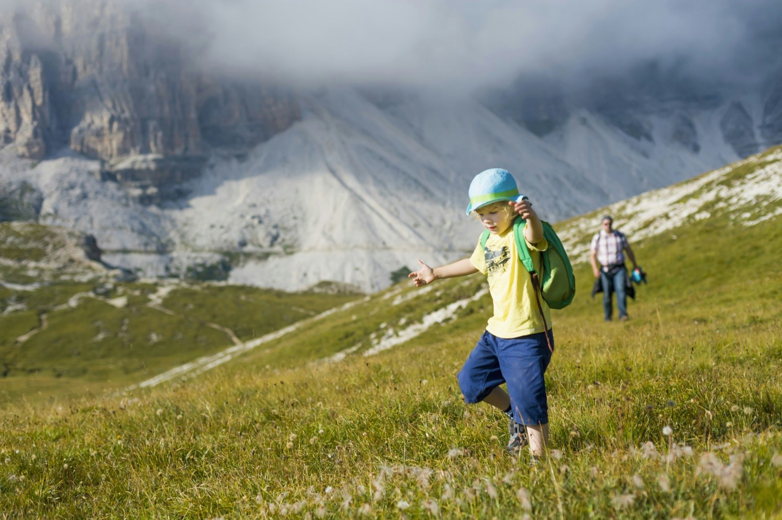 Little boy in a yellow t-shirt and backpack runs through an alpine meadow in the Dolomites with his dad