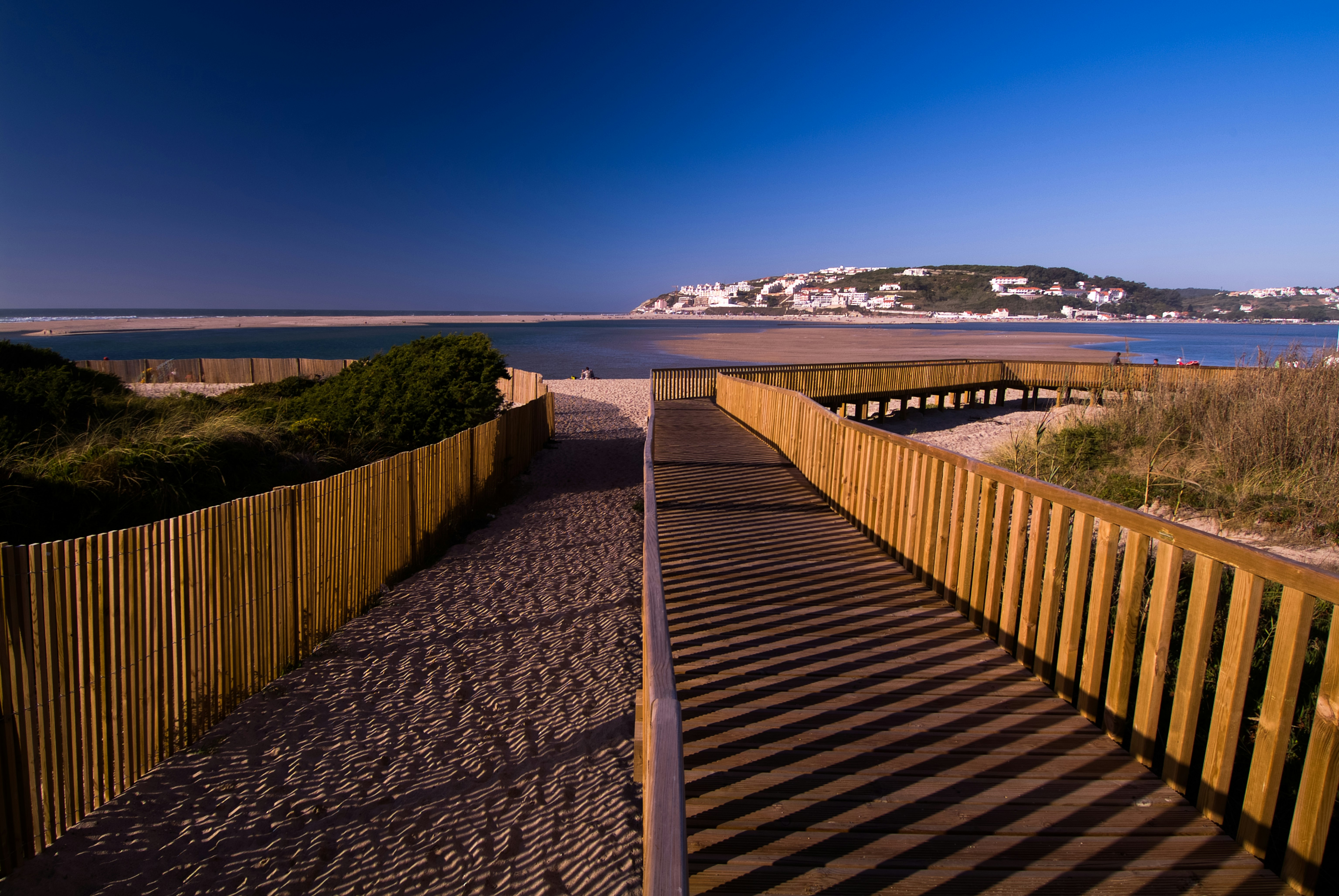 The golden hour sun casts long shadows across a raised boardwalk over sand dunes. The boardwalk stretches ahead, and the sea is is beyond. In the distance is a village with white buildings on a forested hill.