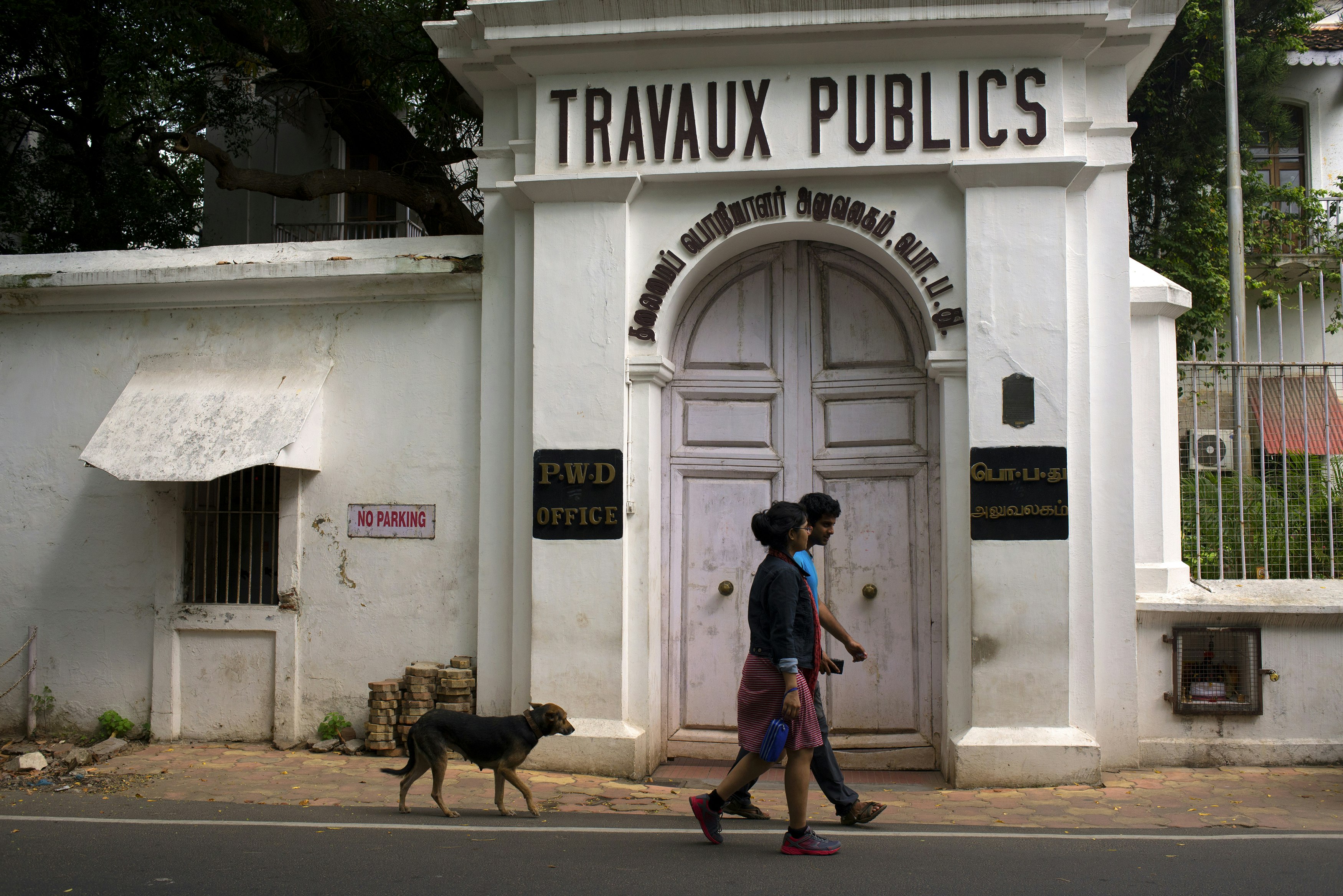 A young Indian couple in modern dress, followed by a dog, walk in front of the white colonial-style Travaux Publics government building