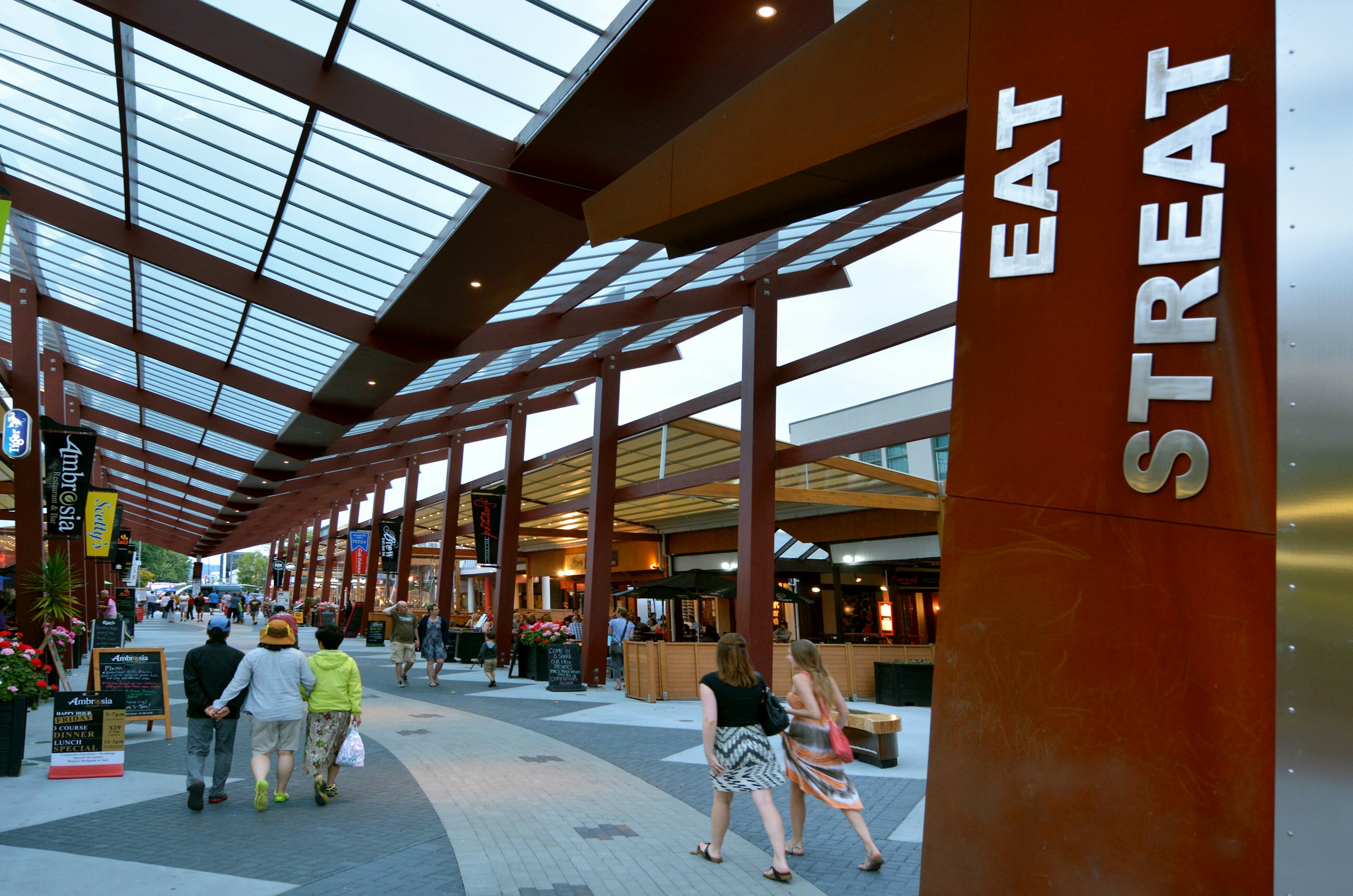 People walk down a paved street under a glass and steel canopy. There are bars and restaurants on either side and in the foreground is a sign that says 'Eat Streat' in capital letters.