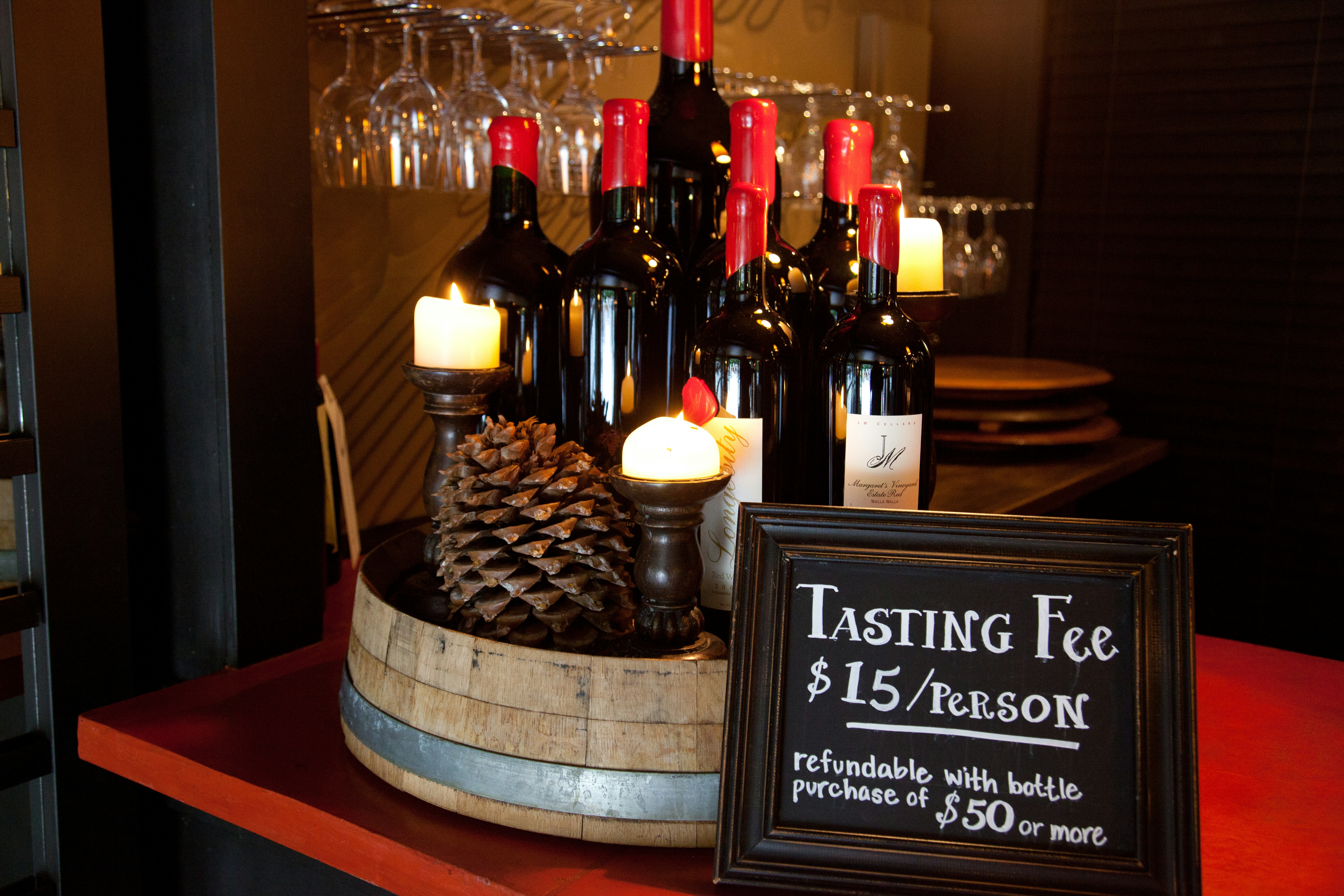 An arrangement of red wine bottles topped with red melted wax, pine cones, and candles sits on a dimly lit red counter with a chalkboard sign that reads "tasting fee $15 per person" in white letters. In the background, clear wine glasses hang upside down from a shelf.