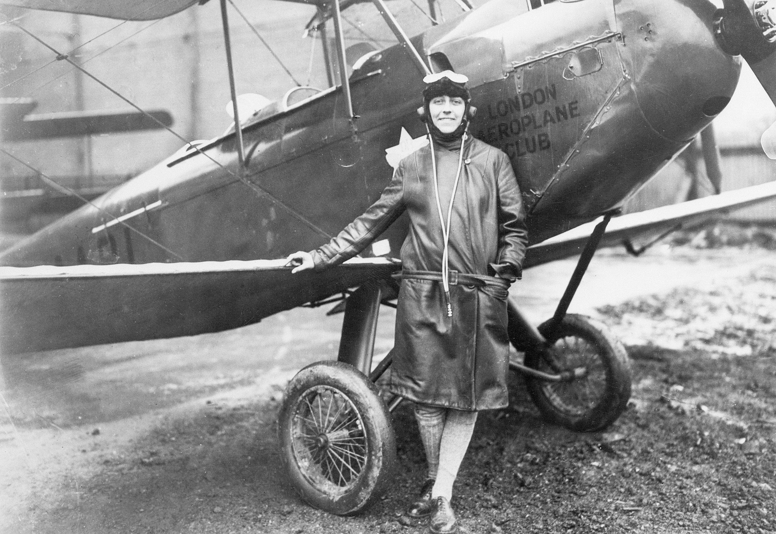 Black-and-white image of a woman wearing a three-quarter length coat with flight goggles on her head standing near a biplane