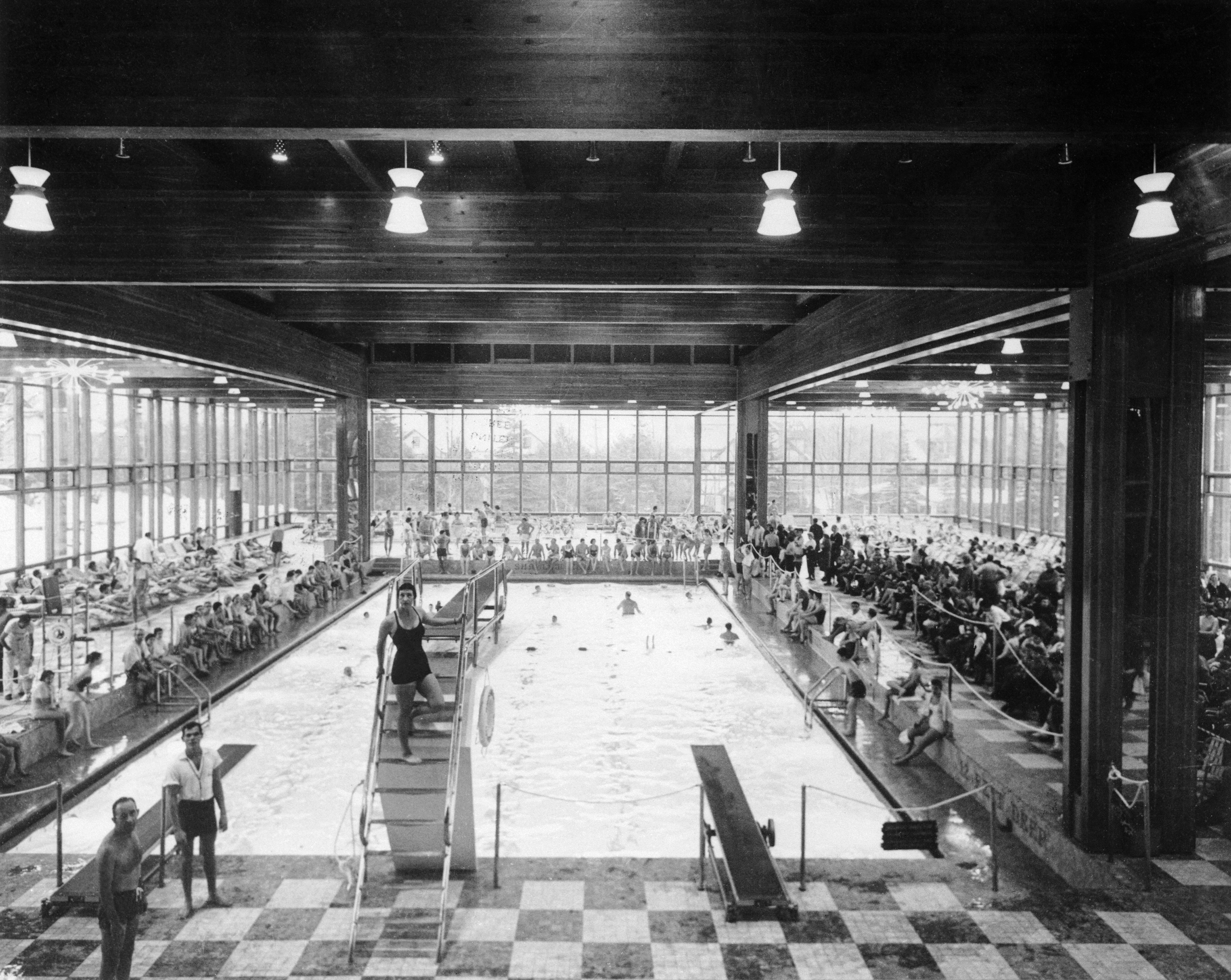 A black and white photo circa 1960 shows a woman in a black one-piece bathing suit and swim cap standing on the stairs to a diving board overlooking an enormous indoor swimming pool at Grossinger's resort in the Catskills. The deck of the pool around her is packed full of vacationers, some sitting upright in chairs just on the edge of the pool and some lounging on deck chairs further back. The cavernous room has glass walls looking out on the grounds and a checkerboard tiled floor.
