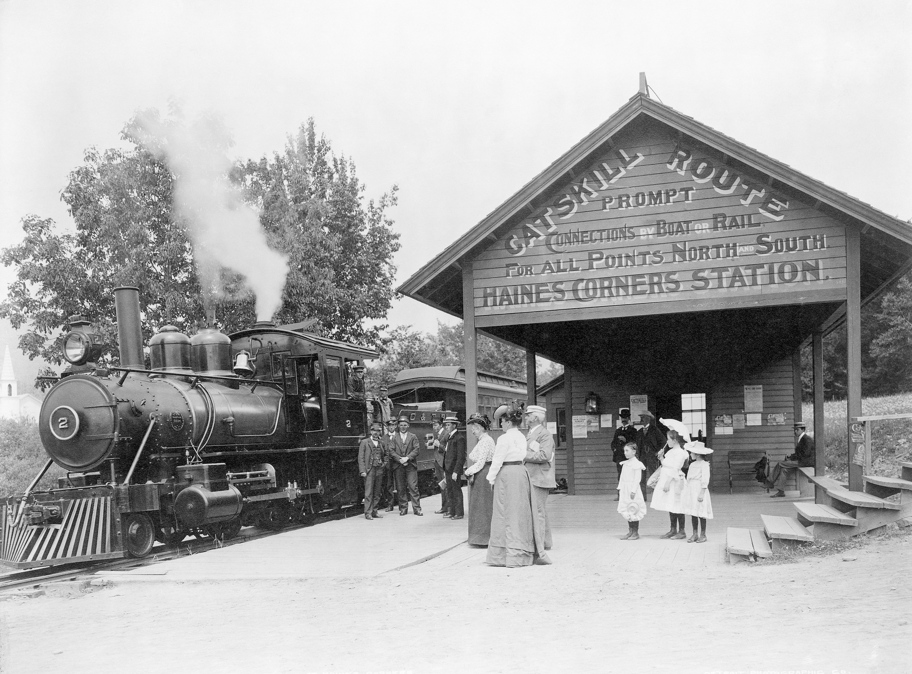 A black and white photo from 1905 shows women and children in long skirts, white shirtwaists, and decorated straw hats standing by a black steam locomotive. Four men in suits and straw brim hats stand by the train, while a group of three little girls in black stockings, white dresses, and large dress hats stand under a wooden siding. Painted on its side gable, facing the viewer, is lettering reading "Catskill Route Prompt Connections by Boat or Rail for all points North and South Haines Corners Station"