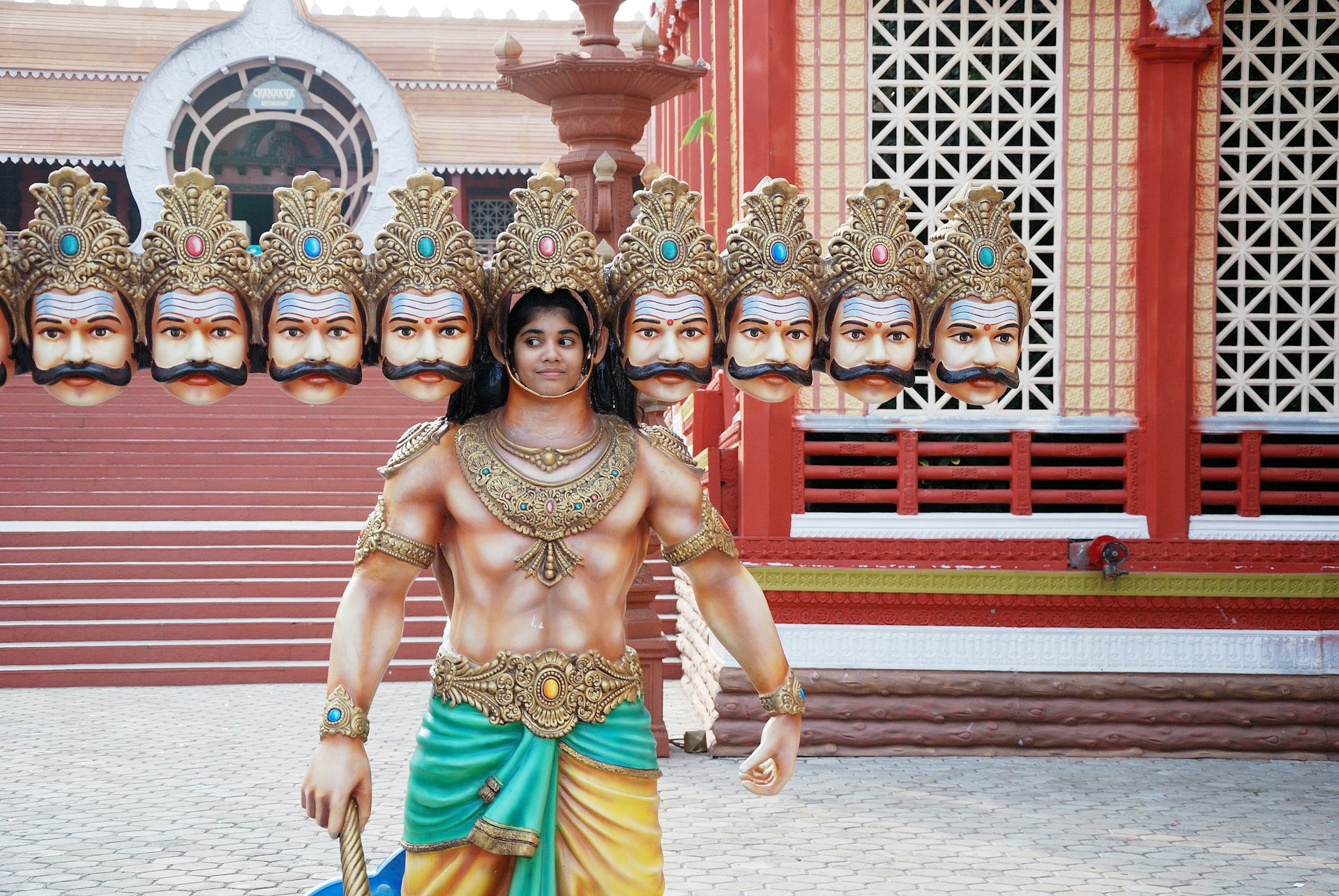 A young Indian girl poses in a cutout of a character with many heads from the film "ten months of Ravana" who is holding a sword and wearing yellow pants and a teal wrap around his waist secured with a brooch