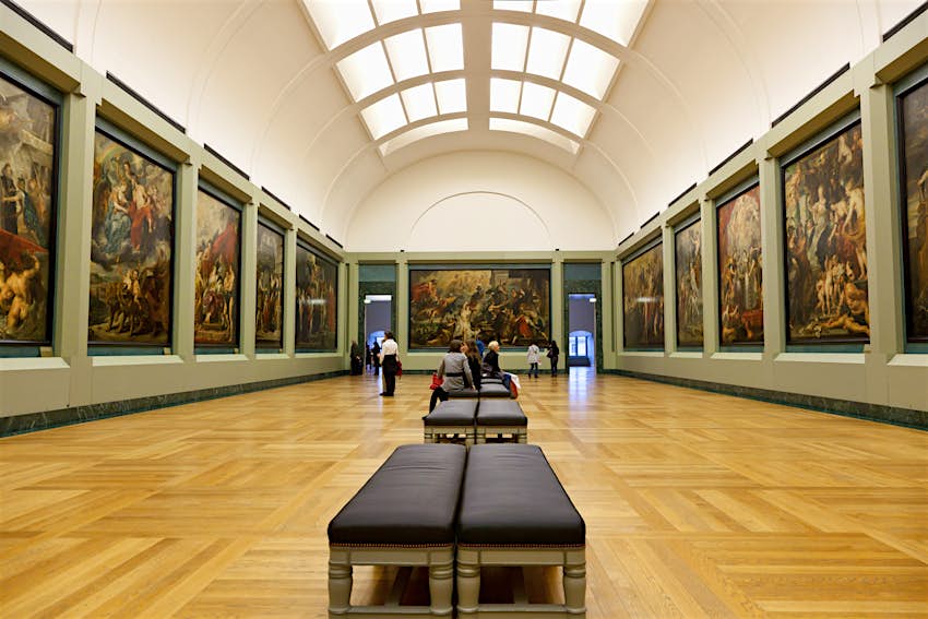 A view of the Galerie Medici: a wooden-floored gallery with a series of wooden benches running down the center of the room.  Large paintings hang on the walls and skylights are installed in the ceiling above.